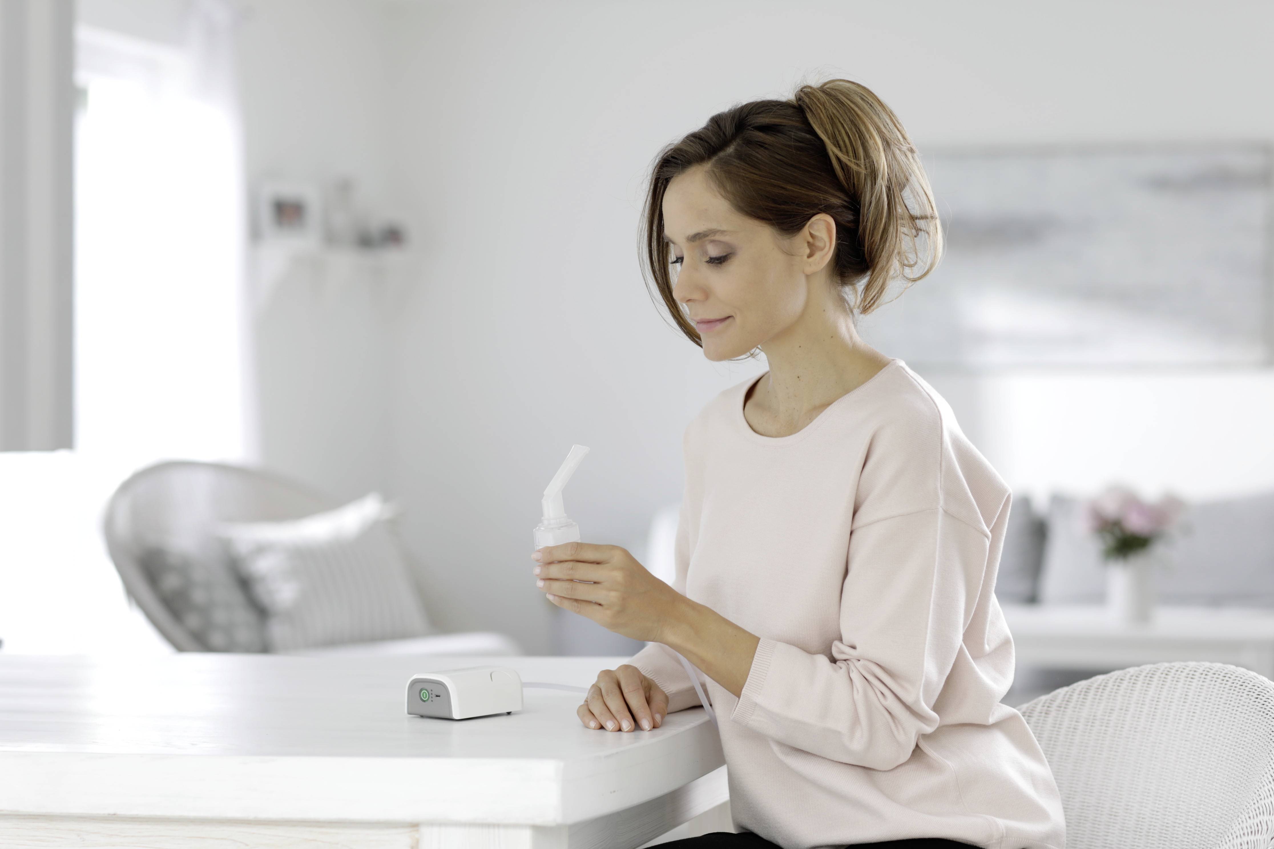 Une femme est assise à une table et tient un appareil d'inhalation. À l'arrière-plan, on peut voir un salon lumineux et un canapé.