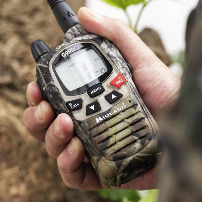 Une main tient un talkie-walkie de camouflage avec un écran affichant plusieurs touches et fonctionnalités.