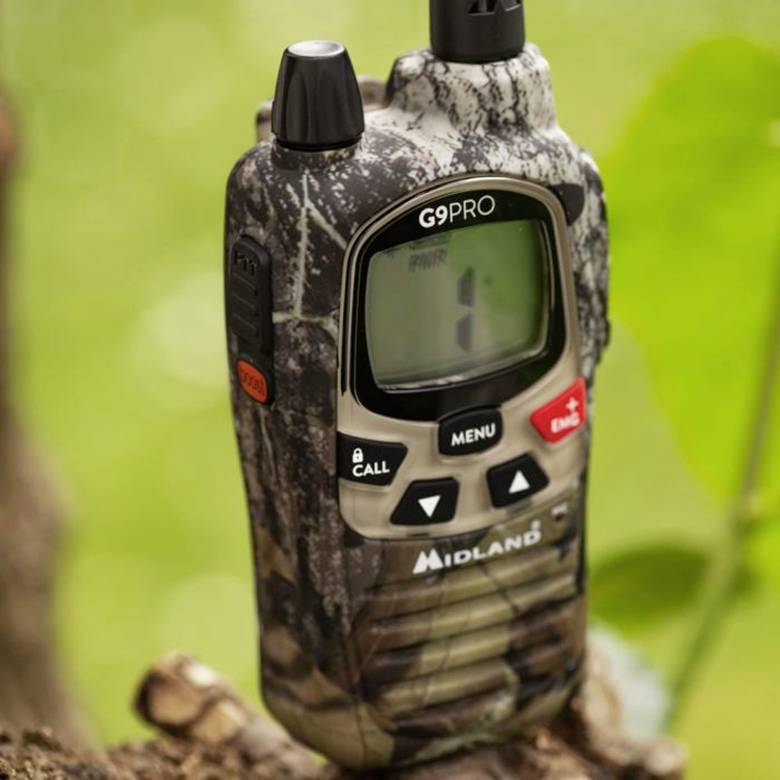Un talkie-walkie de camouflage, modèle Midland G9PRO, est posé sur un tronc d'arbre dans un environnement verdoyant. Son écran est vide.