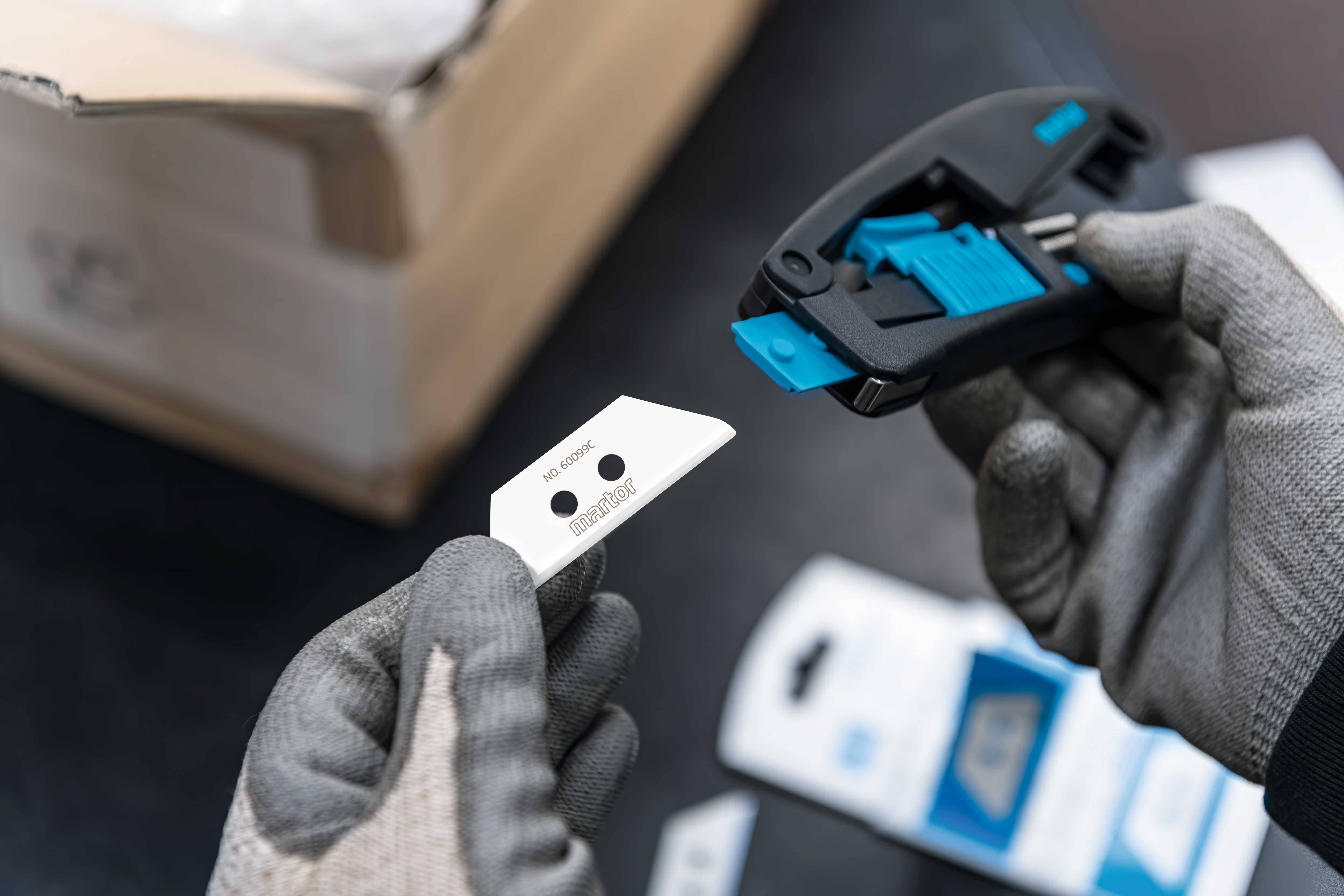 Une personne portant des gants tient un support de lame de rechange et une nouvelle lame, prête à être insérée dans le support. Un carton est visible en arrière-plan.