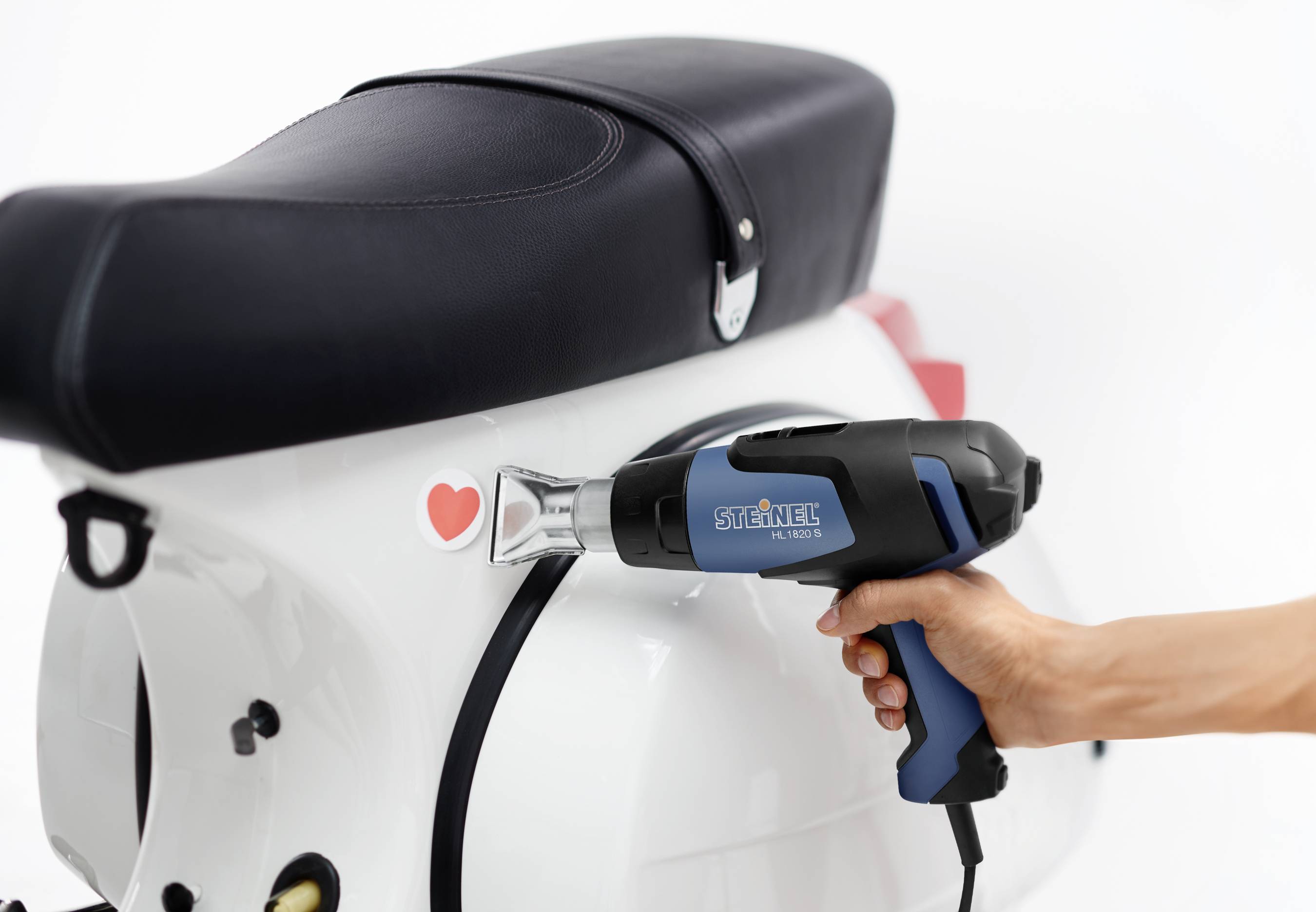 Une main tient un pistolet à air chaud près d'un scooter. Un autocollant en forme de cœur est apposé sur le scooter.