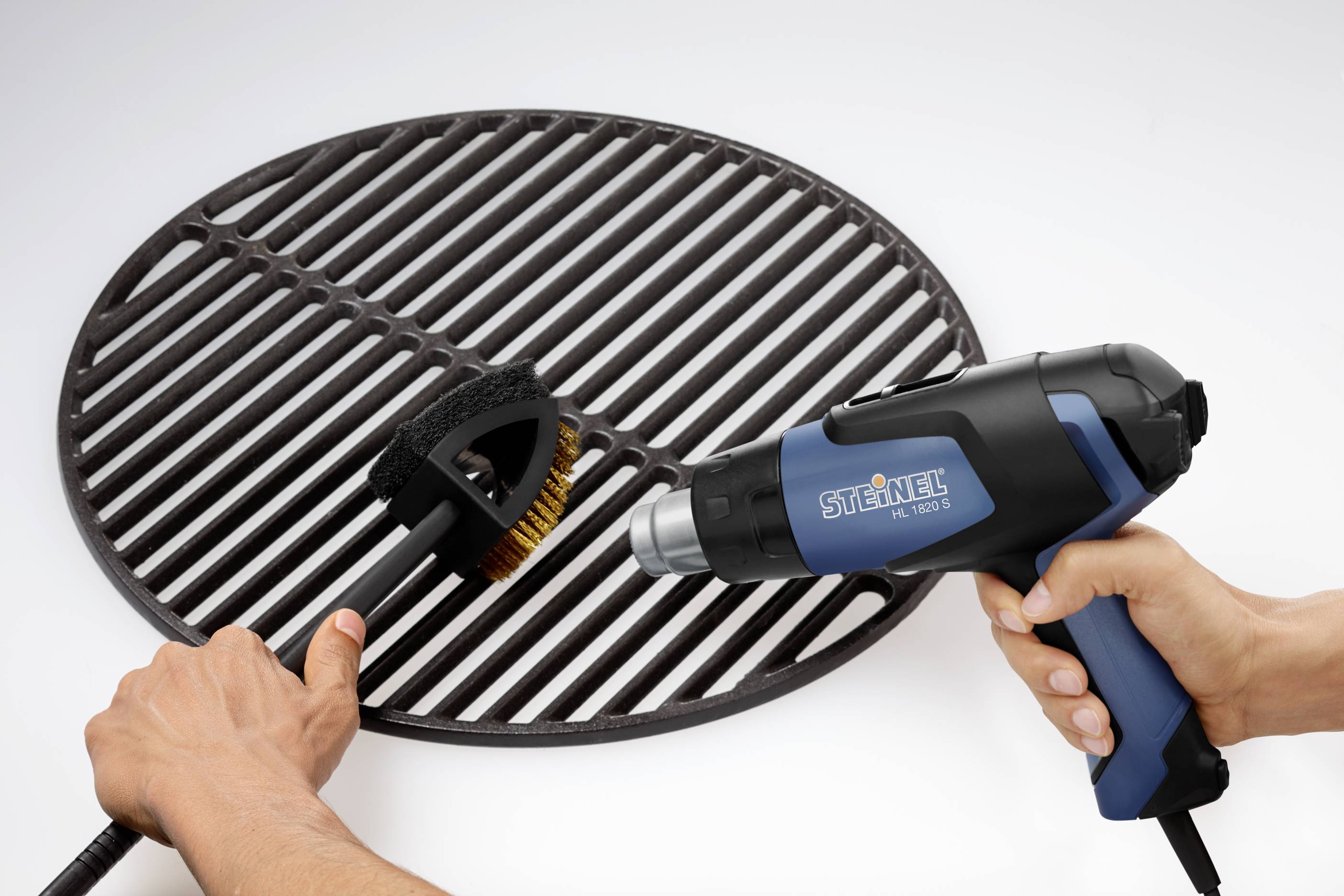 Une personne nettoie une grille de barbecue avec une brosse et un pistolet à air chaud bleu.