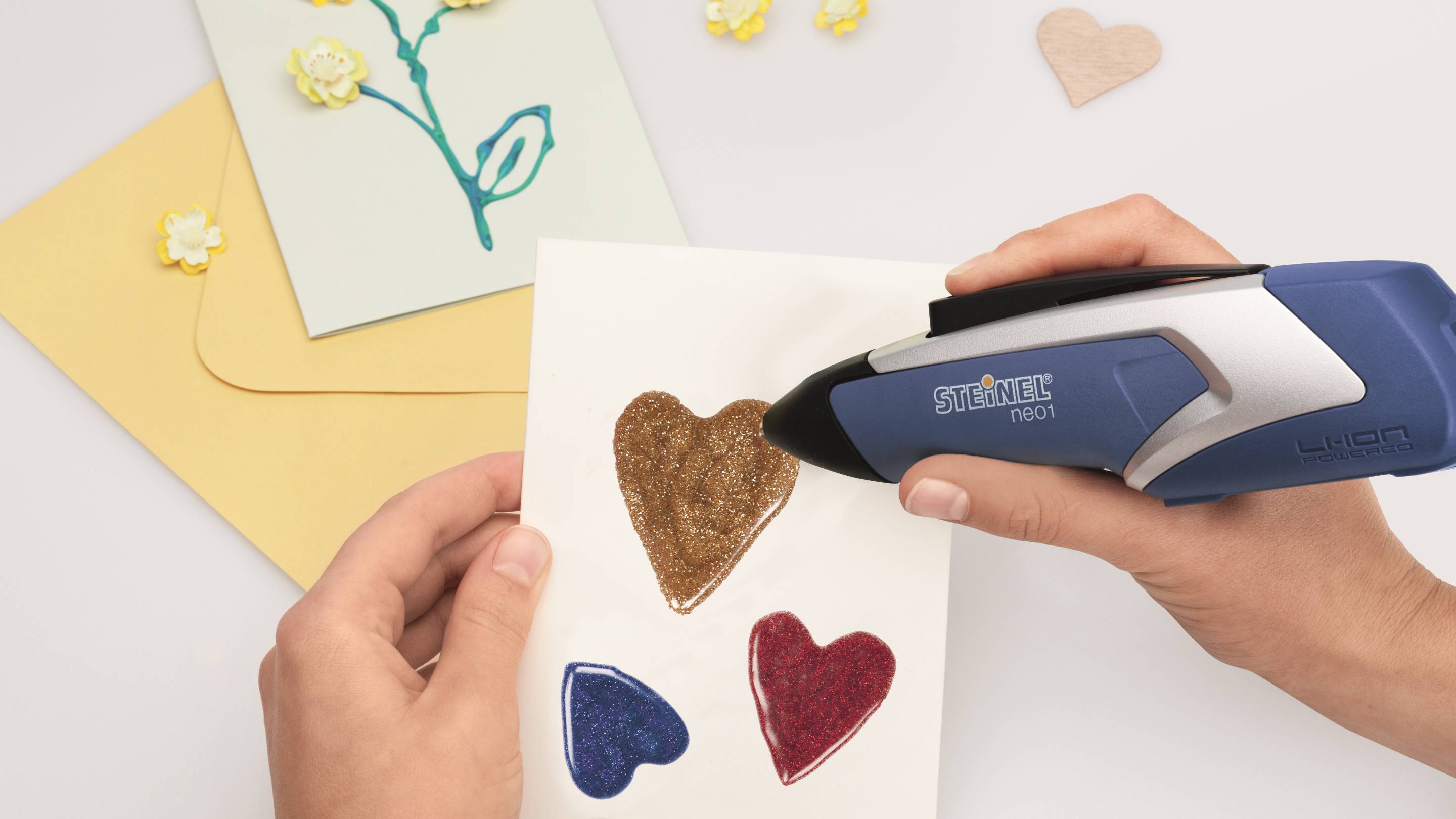 Une personne travaille avec un pistolet à colle chaude bleu sur une carte décorée de cœurs colorés et brillants. À l'arrière-plan, on aperçoit d'autres matériaux de bricolage.