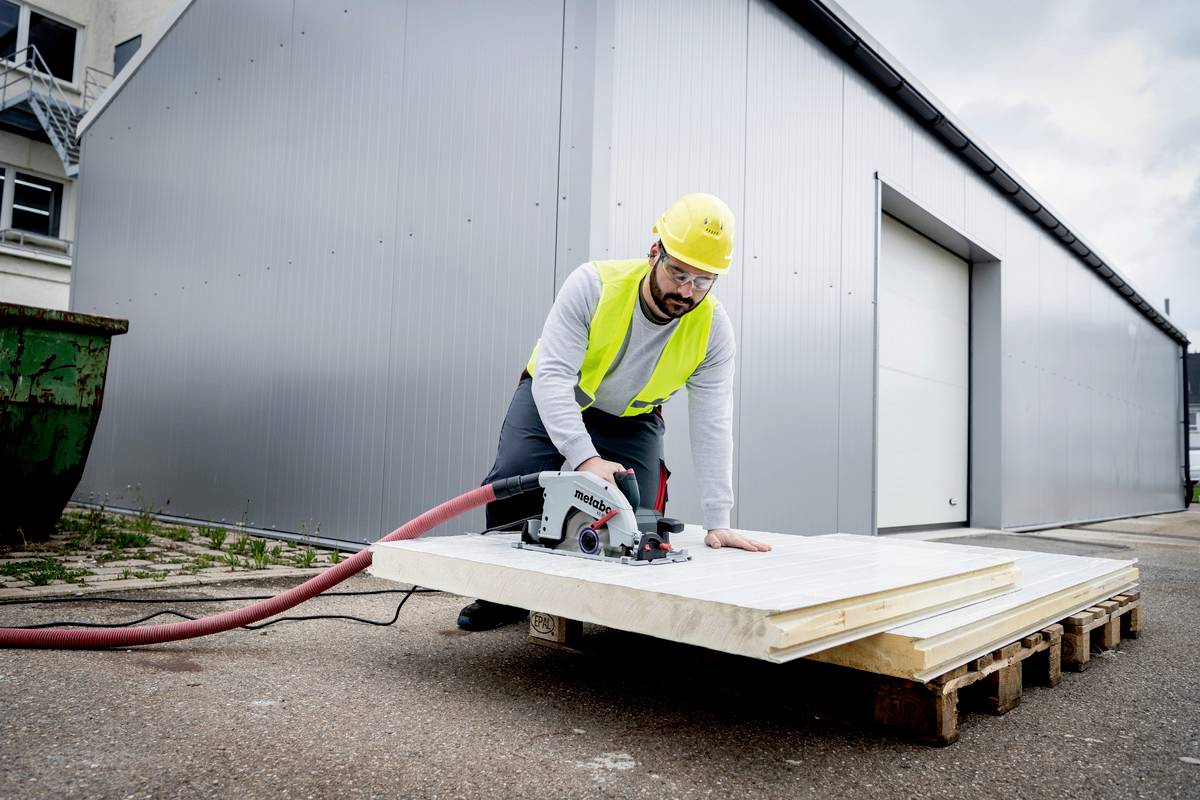 Un ouvrier coupe une planche avec une scie circulaire sur un site industriel, portant un casque, un gilet de sécurité et des gants.