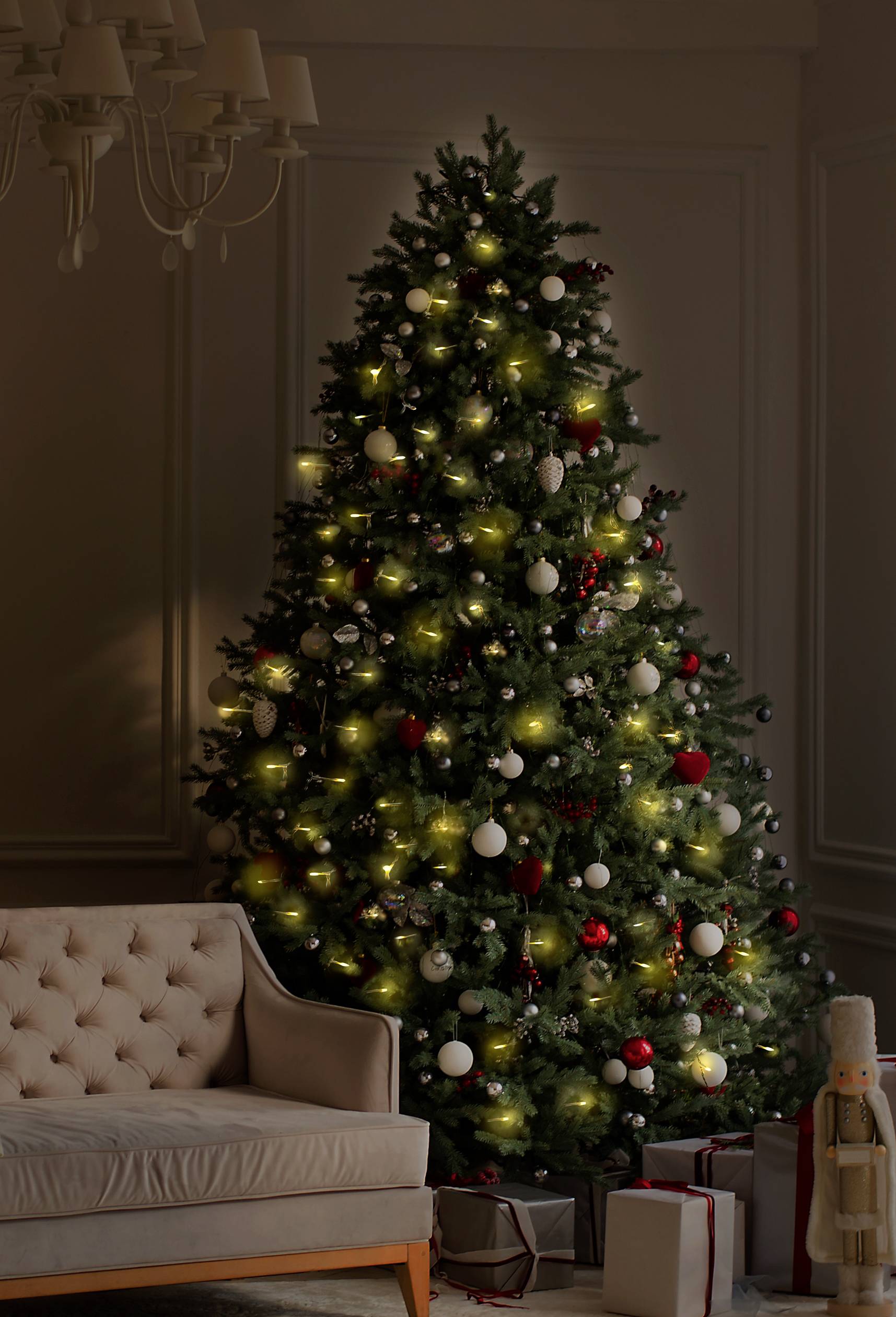 Un sapin de Noël décoré se trouve dans un salon. Il est orné de boules rouges et blanches et de lumières, avec des cadeaux disposés en dessous.