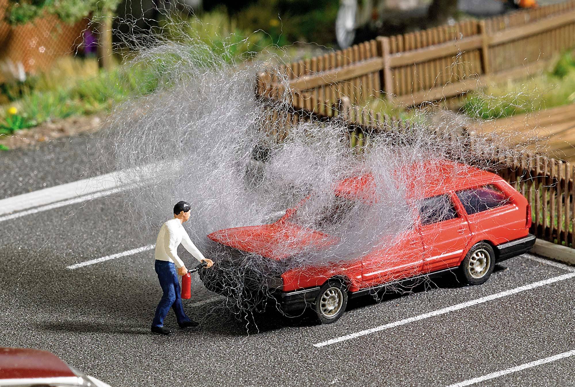 Un homme tient un extincteur et se tient devant une voiture rouge sur un parking, dont le compartiment moteur dégage de la fumée.