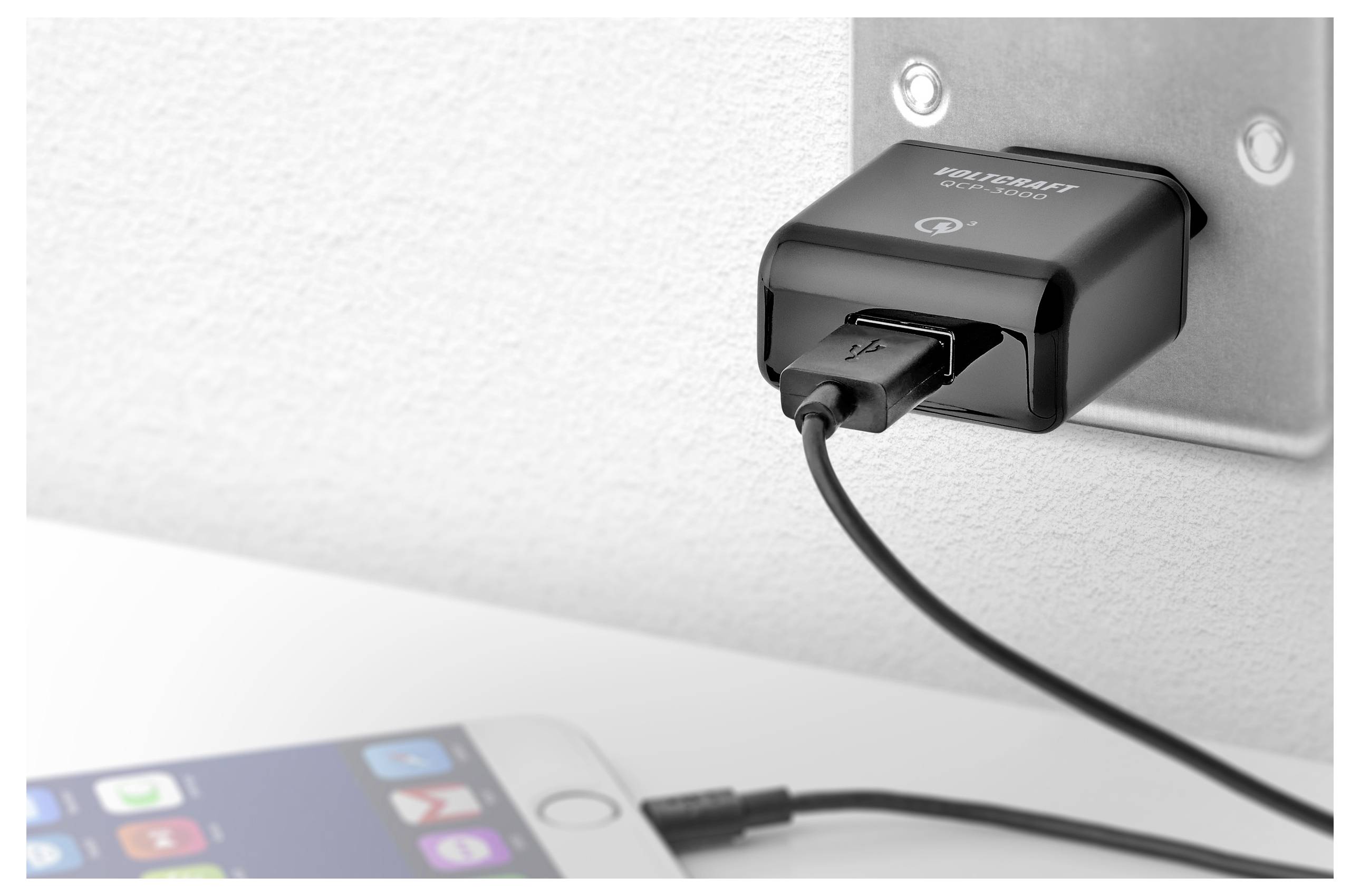 Un chargeur USB noir branché dans une prise murale charge un smartphone sur une table blanche au premier plan.