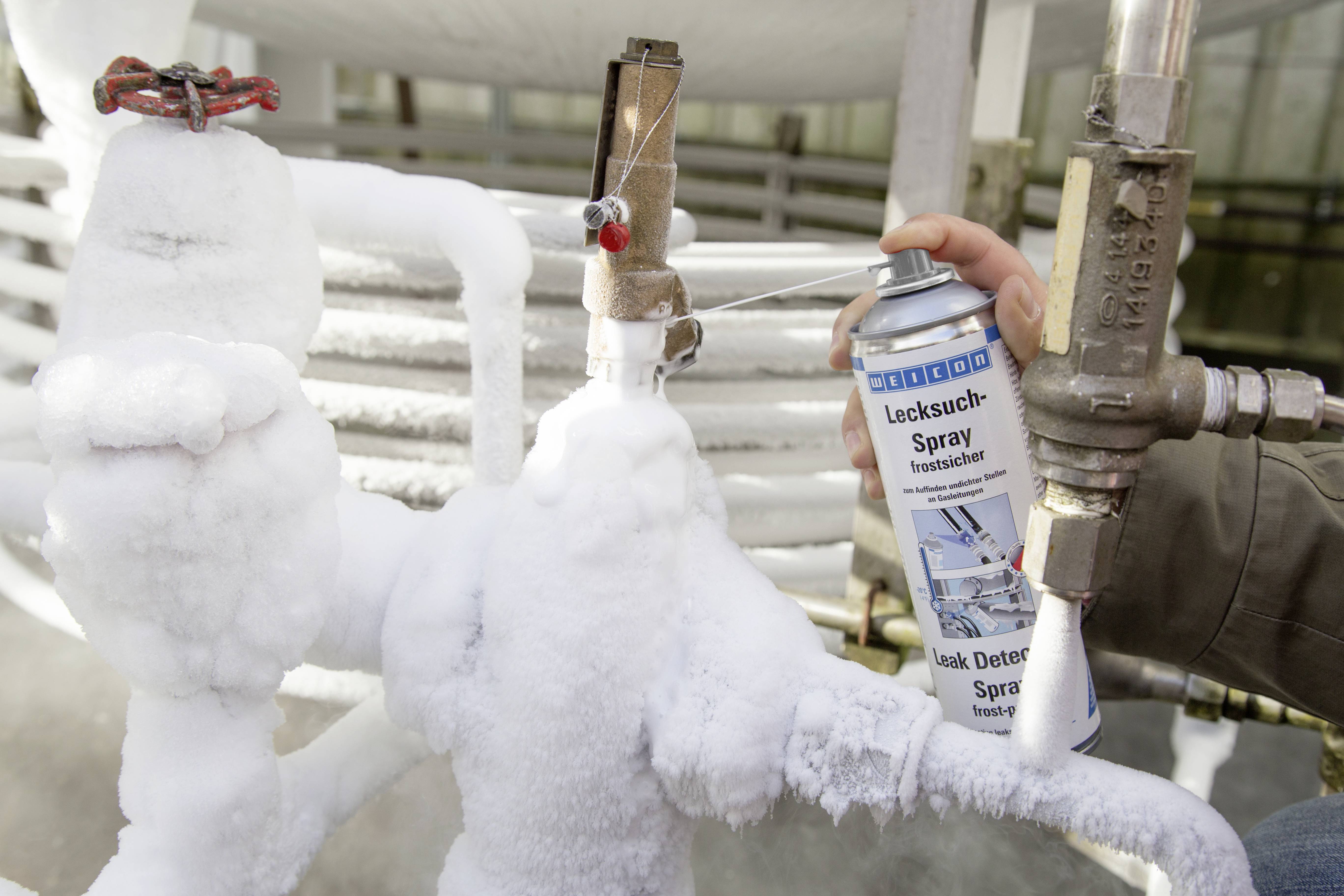 Une personne pulvérise un détecteur de fuite sur des tuyaux gelés afin de localiser les fuites. Les tuyaux sont fortement recouverts de glace.