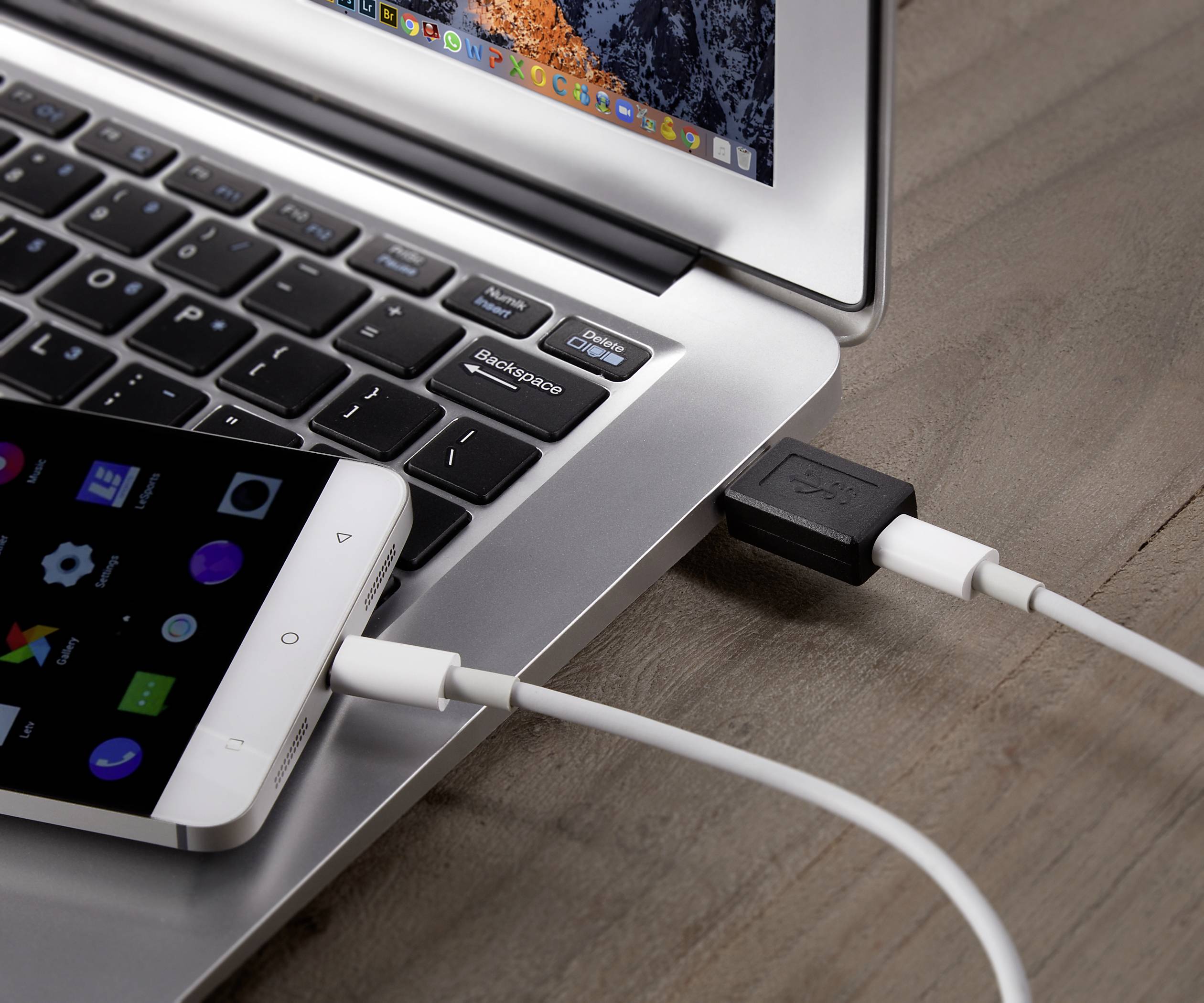 Smartphone connecté à un ordinateur portable via un câble USB, visible sur une table en bois. L'écran de l'ordinateur portable affiche des icônes colorées.