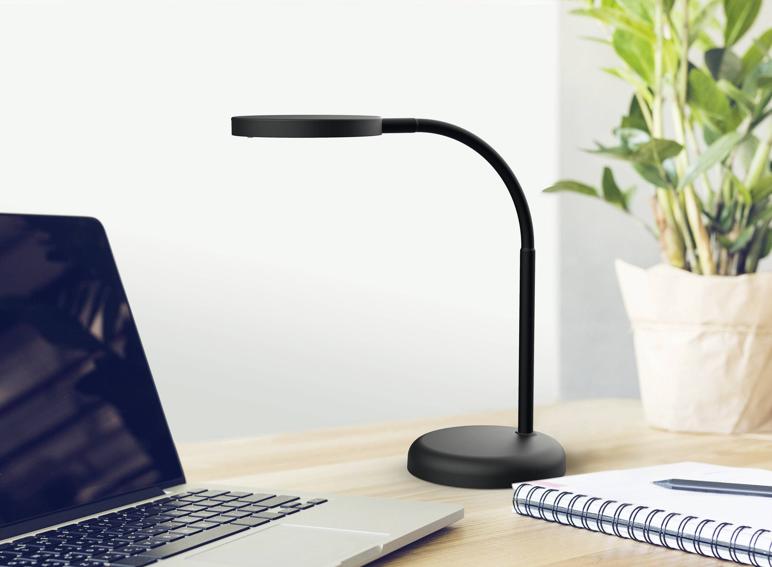 Une lampe de bureau noire est posée sur une table à côté d'un ordinateur portable et d'un carnet. Une plante est visible en arrière-plan.