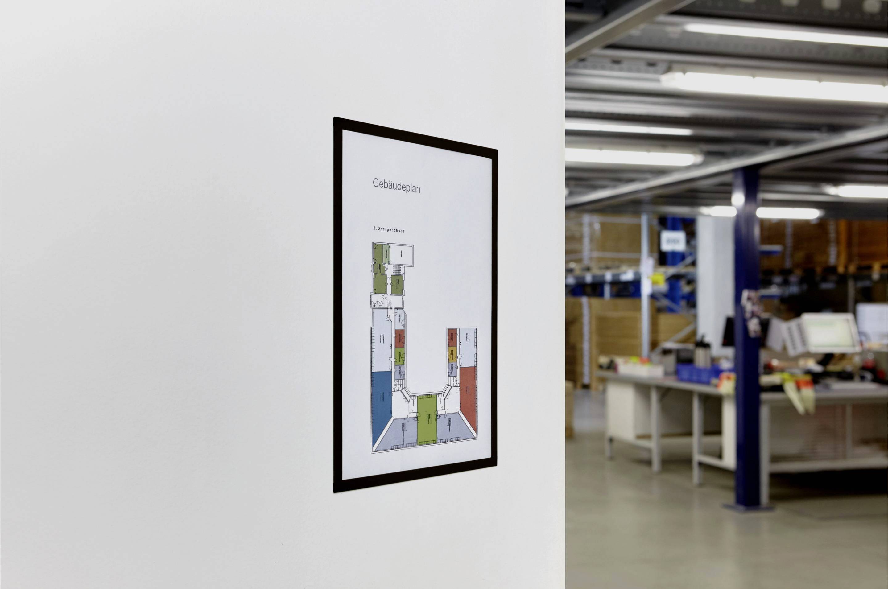 Sur un pan de mur blanc est accroché un plan de bâtiment, avec en arrière-plan un espace de bureau équipé de bureaux et d'étagères.