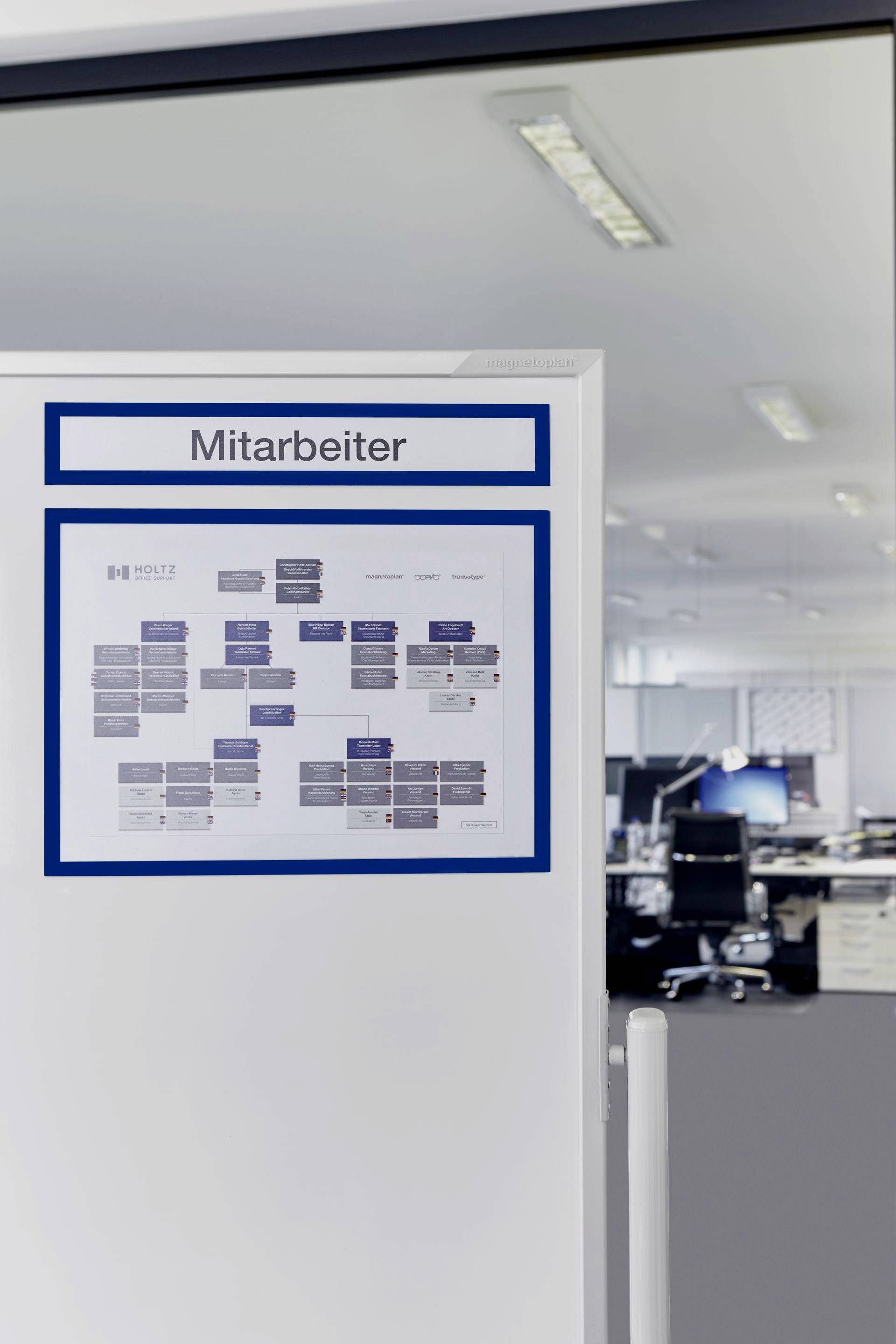 Un bureau avec plusieurs bureaux et ordinateurs ; au premier plan, un tableau d'affichage portant un organigramme sous une étiquette 'Personnel'.