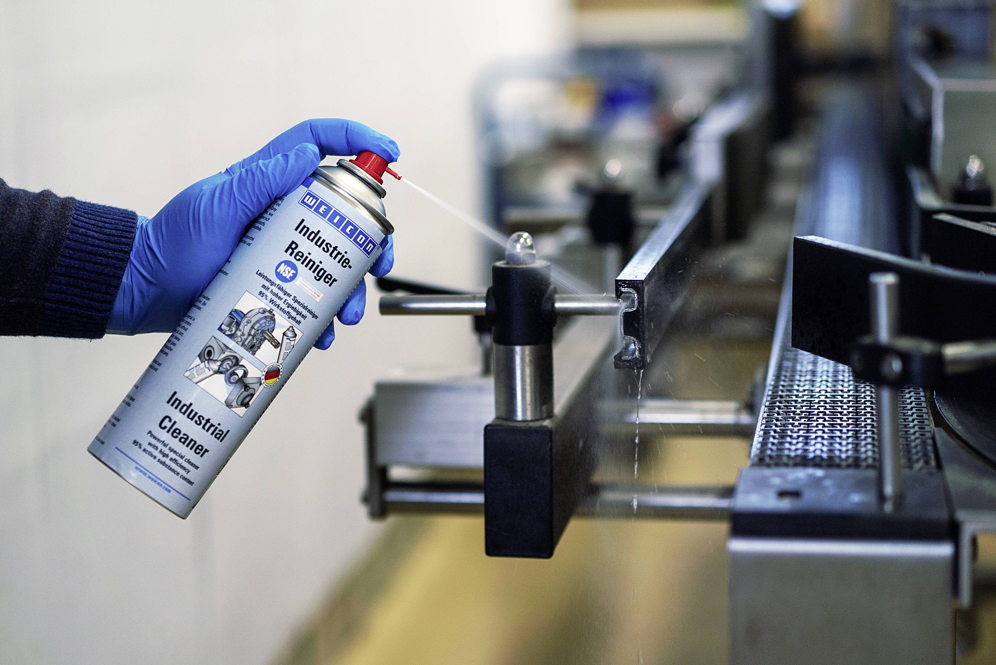 'Une personne portant des gants bleus vaporise un produit de nettoyage industriel d'une bombe aérosol sur une machine en métal pour l'entretien ou le nettoyage.'