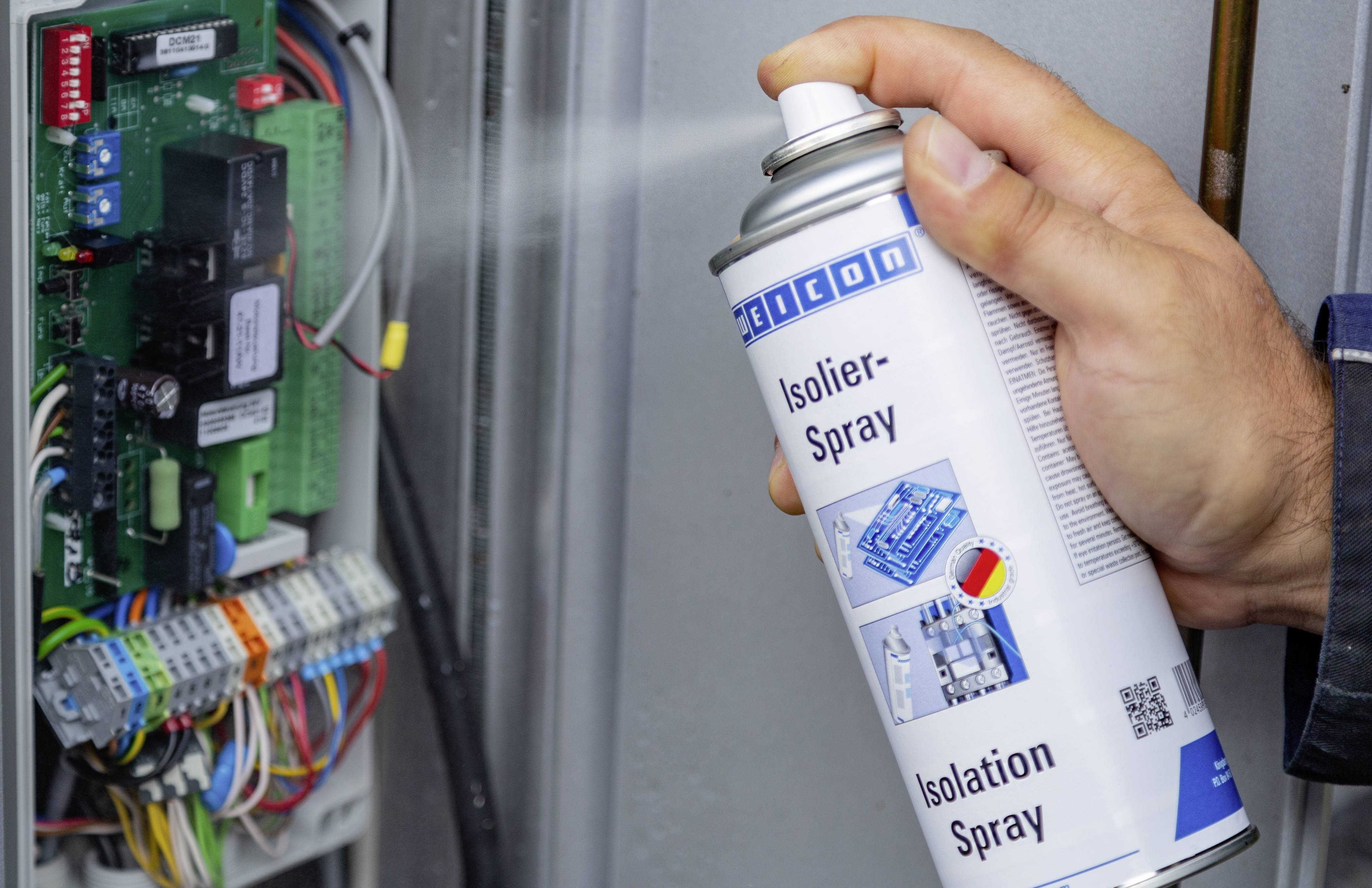 Une personne pulvérise un spray isolant sur un circuit électrique ouvert, afin de le protéger, avec des câbles et des composants visibles.