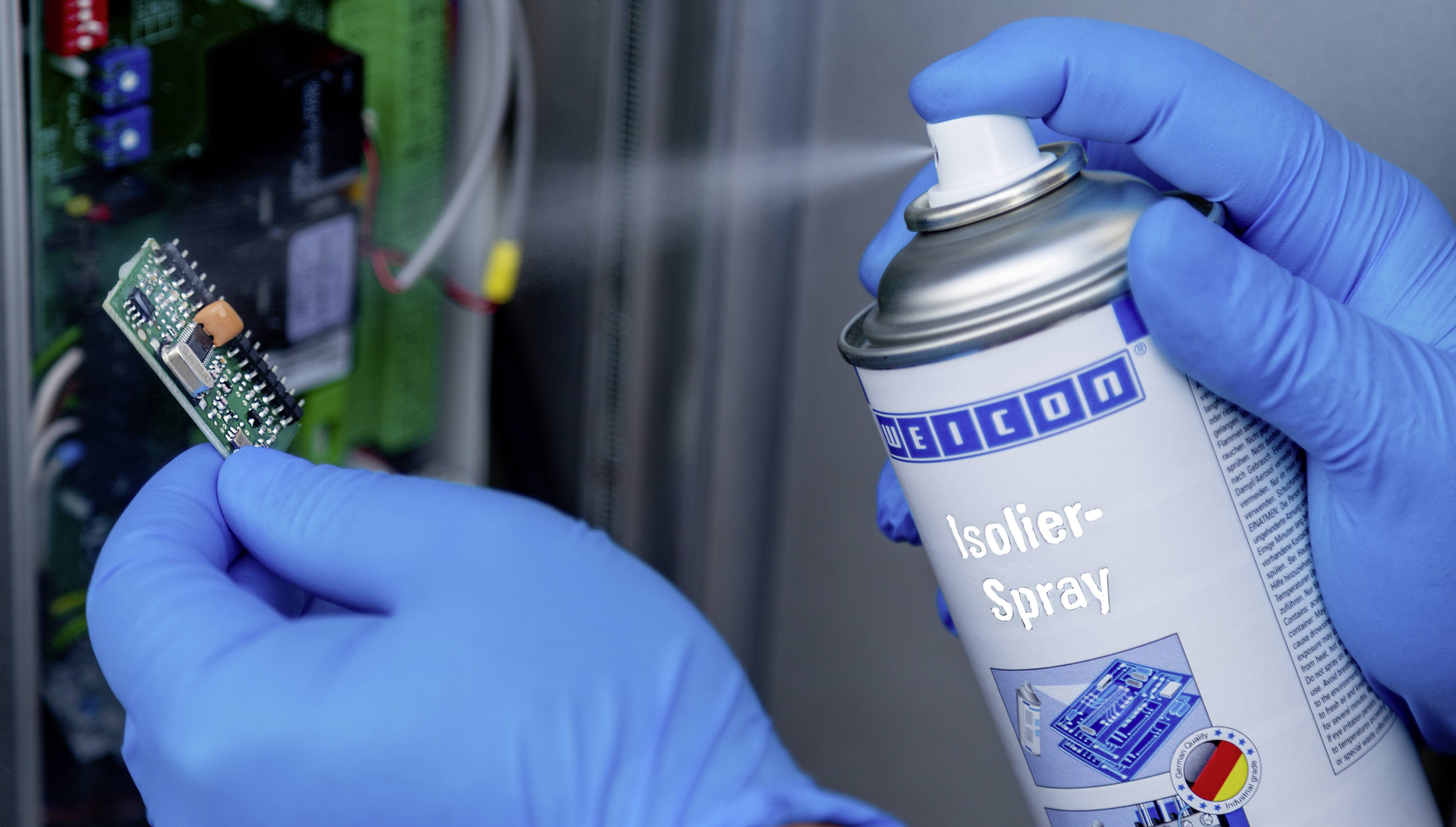 Une personne portant des gants bleus pulvérise un aérosol isolant sur une petite carte électronique.