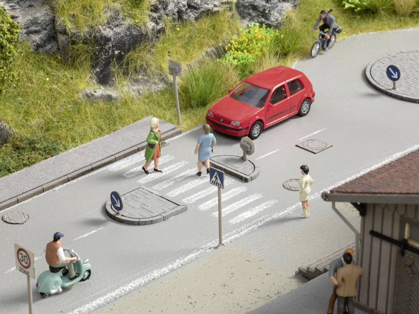 Scène en miniature sur une rue : Deux personnes sur un passage piéton, une voiture rouge s'arrête, un cycliste et un conducteur de scooter sur la chaussée.