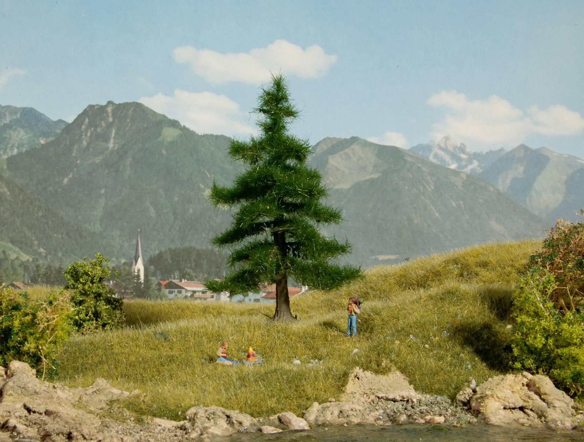 Un paysage idyllique avec un conifère au premier plan, un village avec une église au centre et une chaîne de montagnes en arrière-plan.