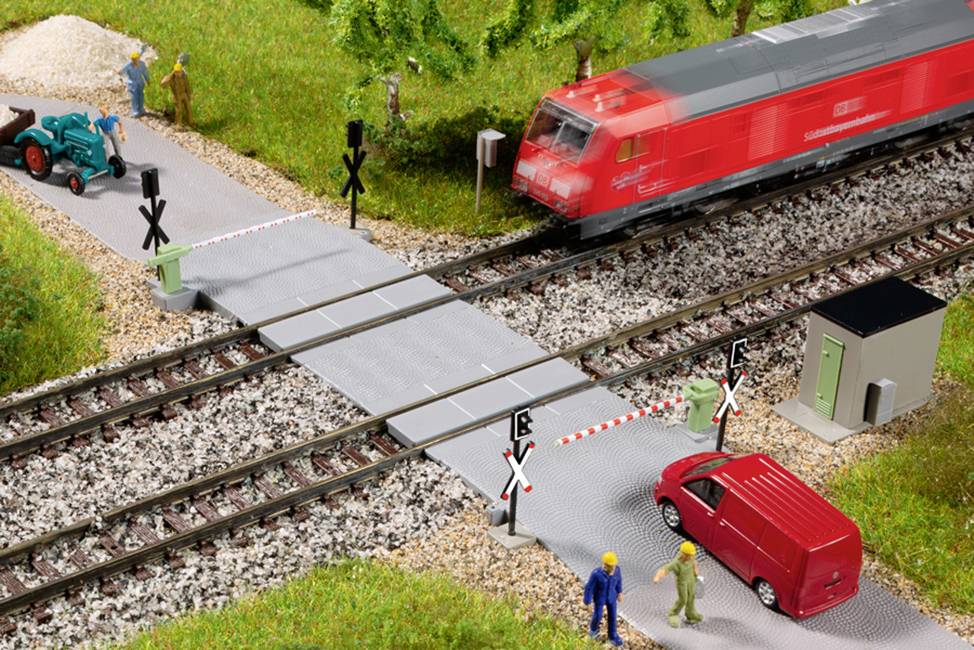Scène de modélisme ferroviaire avec un passage à niveau : Un train rouge traverse la route, une voiture rouge attend. Des ouvriers portent des casques de sécurité.