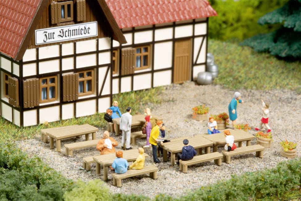 Une maquette miniature représente une maison à colombages allemande avec un panneau indiquant 'À la forge'. Au premier plan, des personnages sont assis sur des bancs et des tables en bois à l'extérieur.