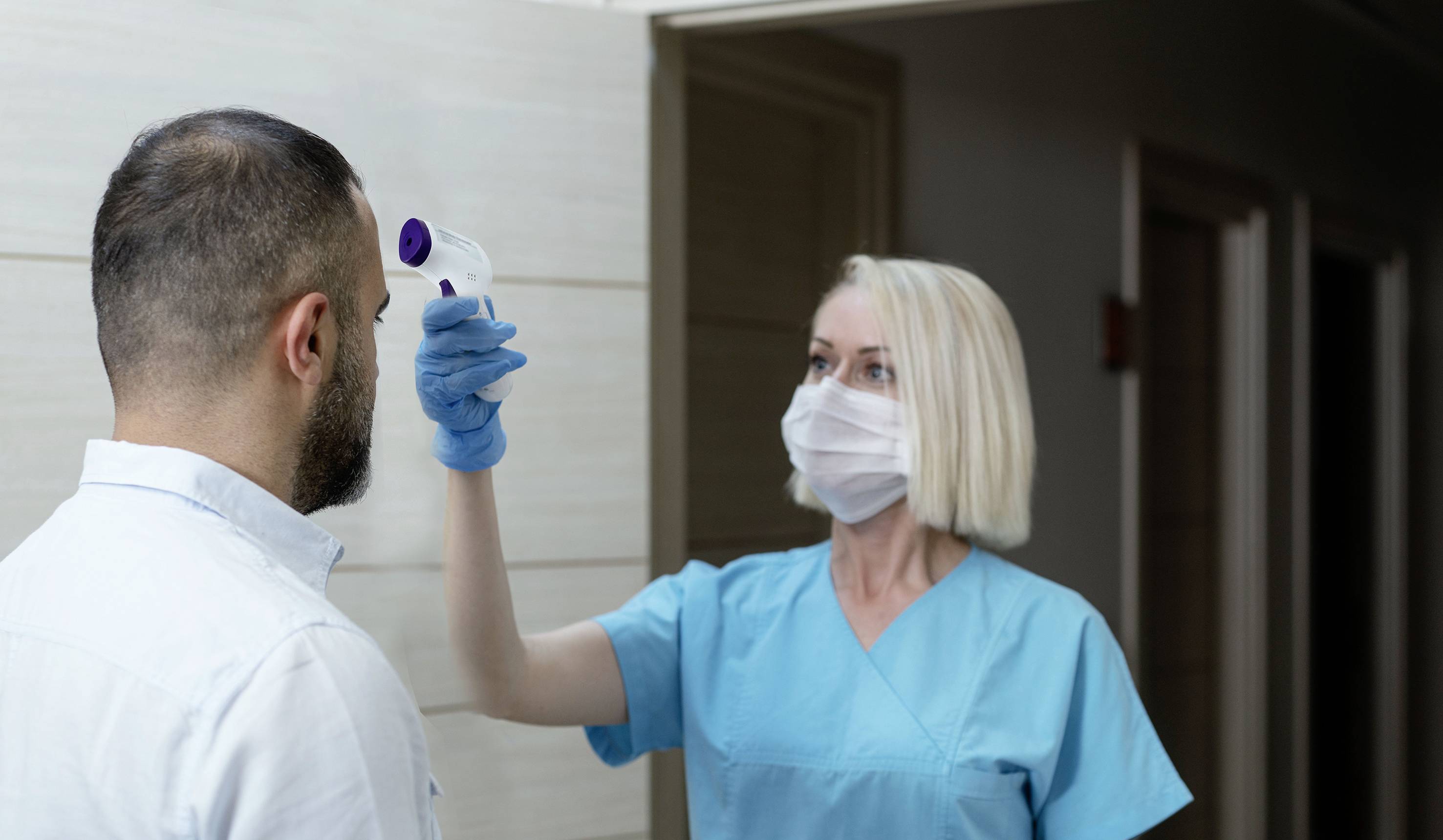 Une infirmière mesure la température d'un homme à l'aide d'un thermomètre frontal. Ils portent tous deux des masques de protection.