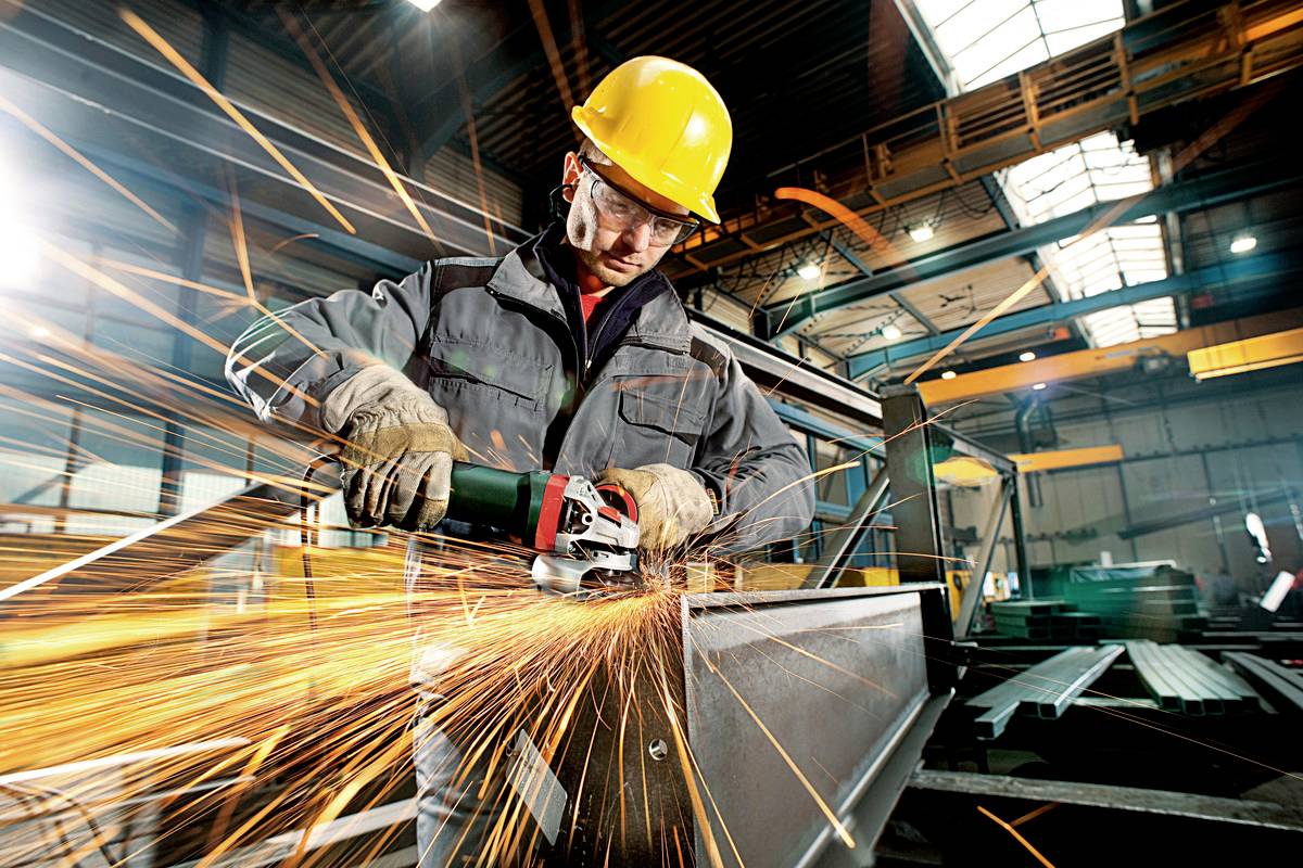 Un ouvrier en tenue de protection meule du métal avec une meuleuse d'angle, faisant jaillir des étincelles dans un environnement industriel.