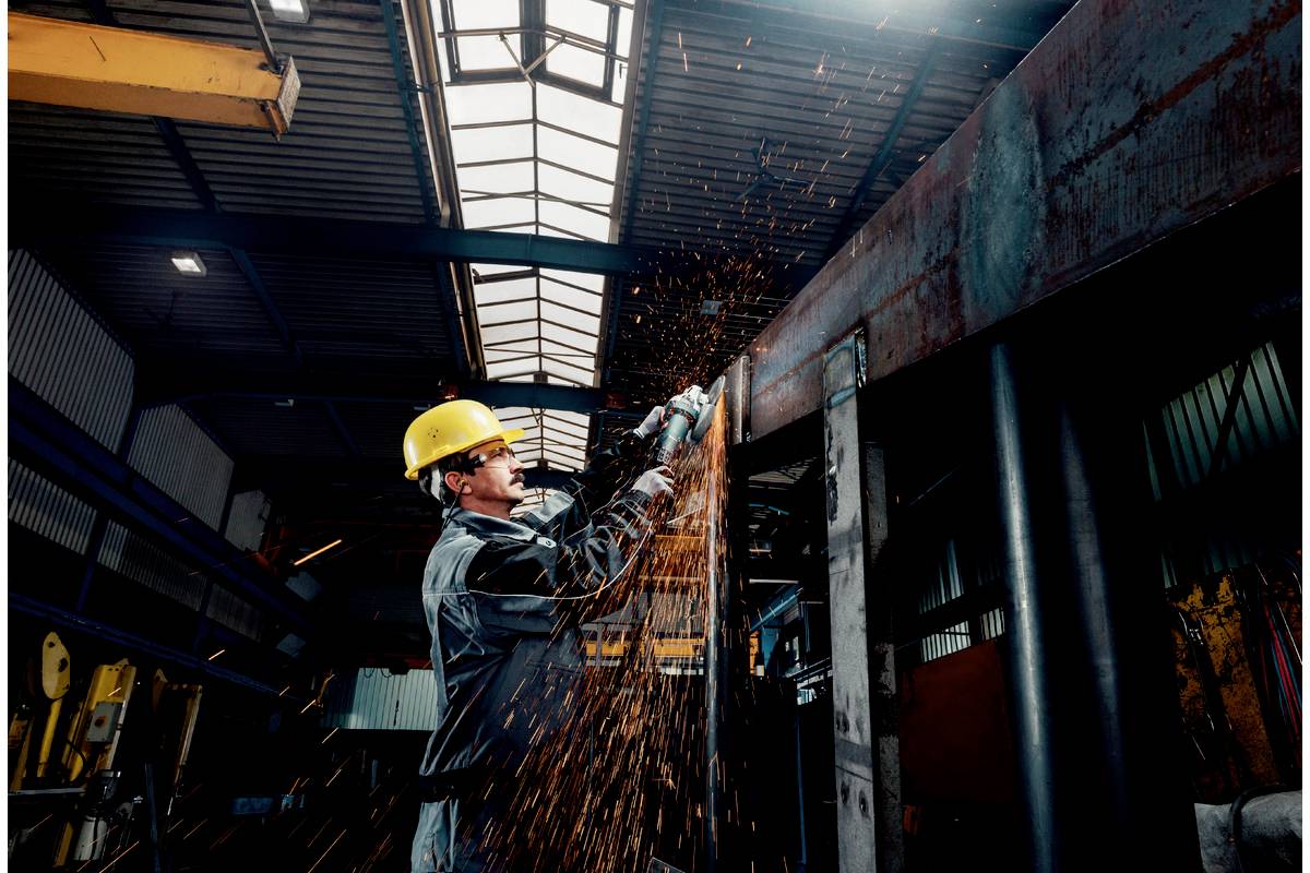 Un ouvrier portant un casque de protection coupe du métal avec une scie électrique dans un hall d'usine. Des étincelles jaillissent dans toutes les directions.