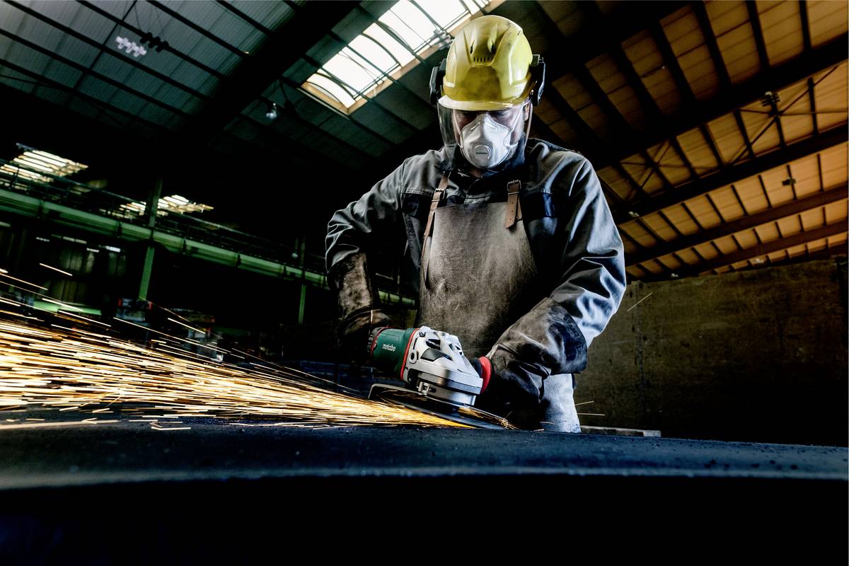 Un ouvrier en tenue de protection et casque meule du métal avec une meuleuse d'angle dans un hall d'usine, avec des gerbes d'étincelles.