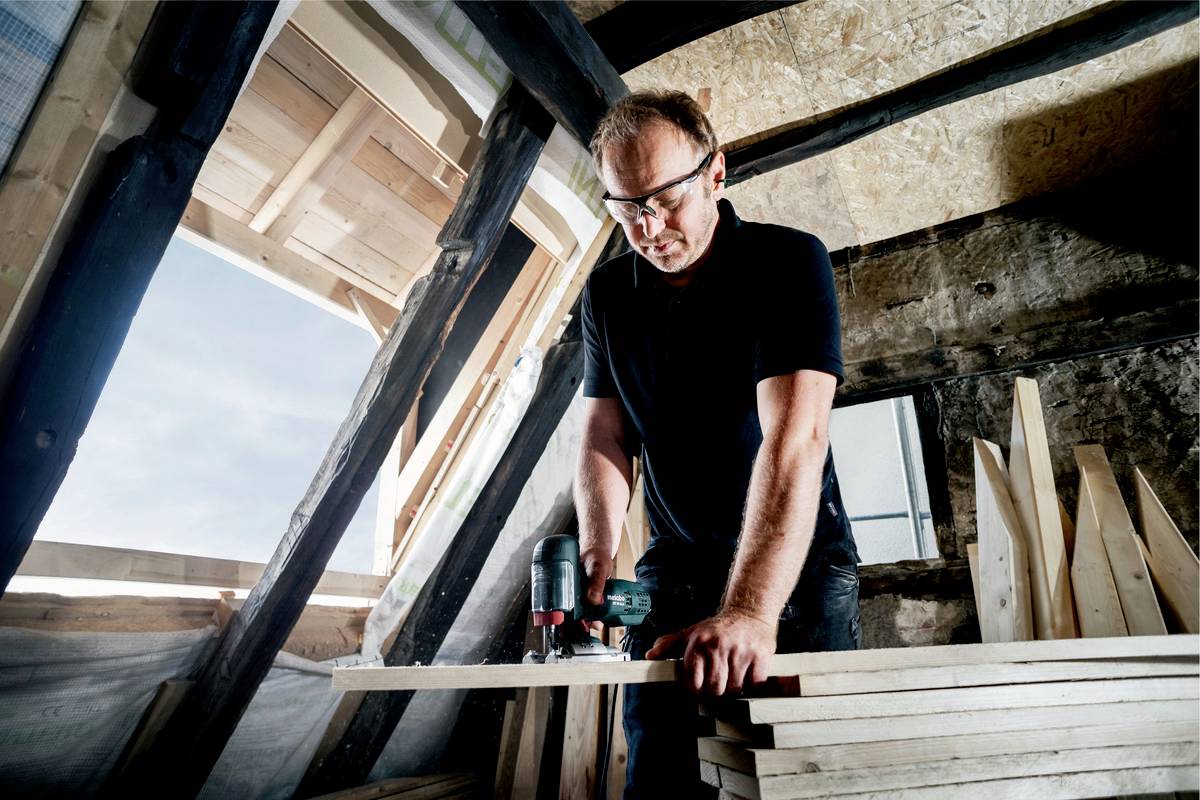 Un artisan travaille dans un grenier, travaillant des panneaux de bois avec un outil électrique, tandis que la lumière du soleil entre par une fenêtre.