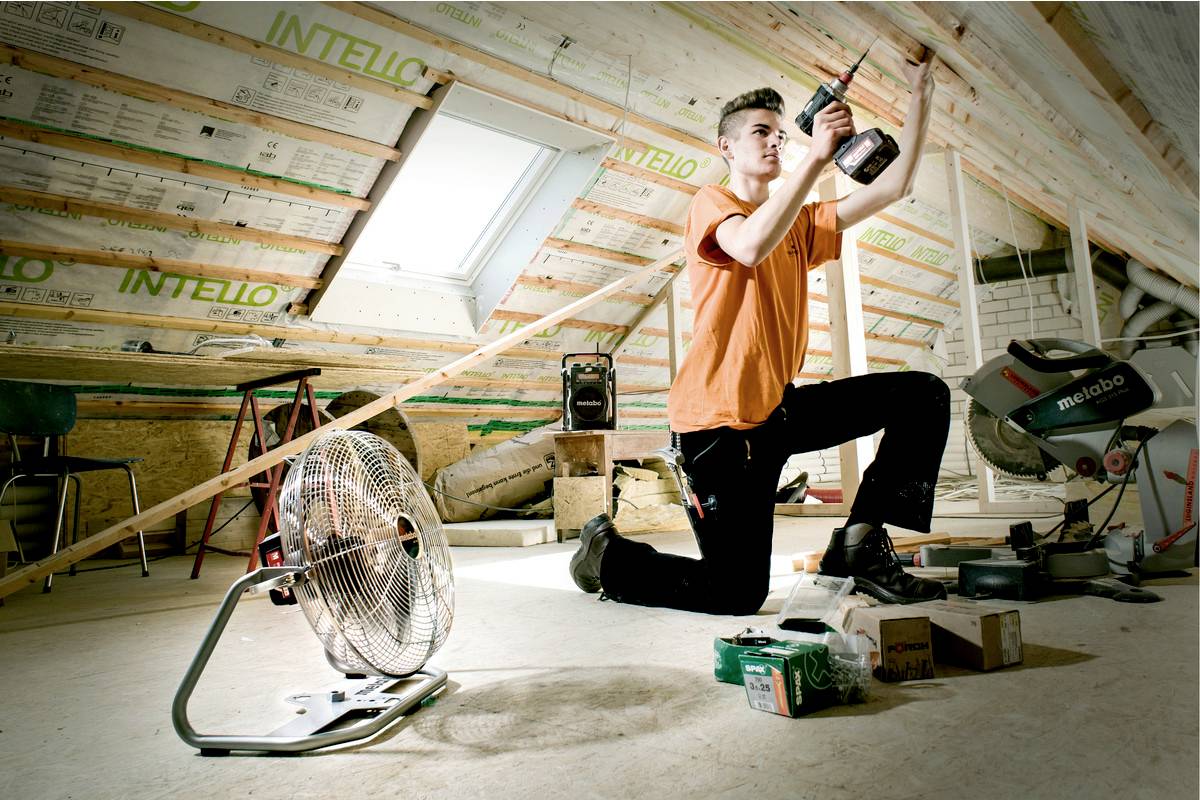 Un artisan est agenouillé dans un grenier et travaille avec un outil électrique. Un ventilateur est placé au premier plan.