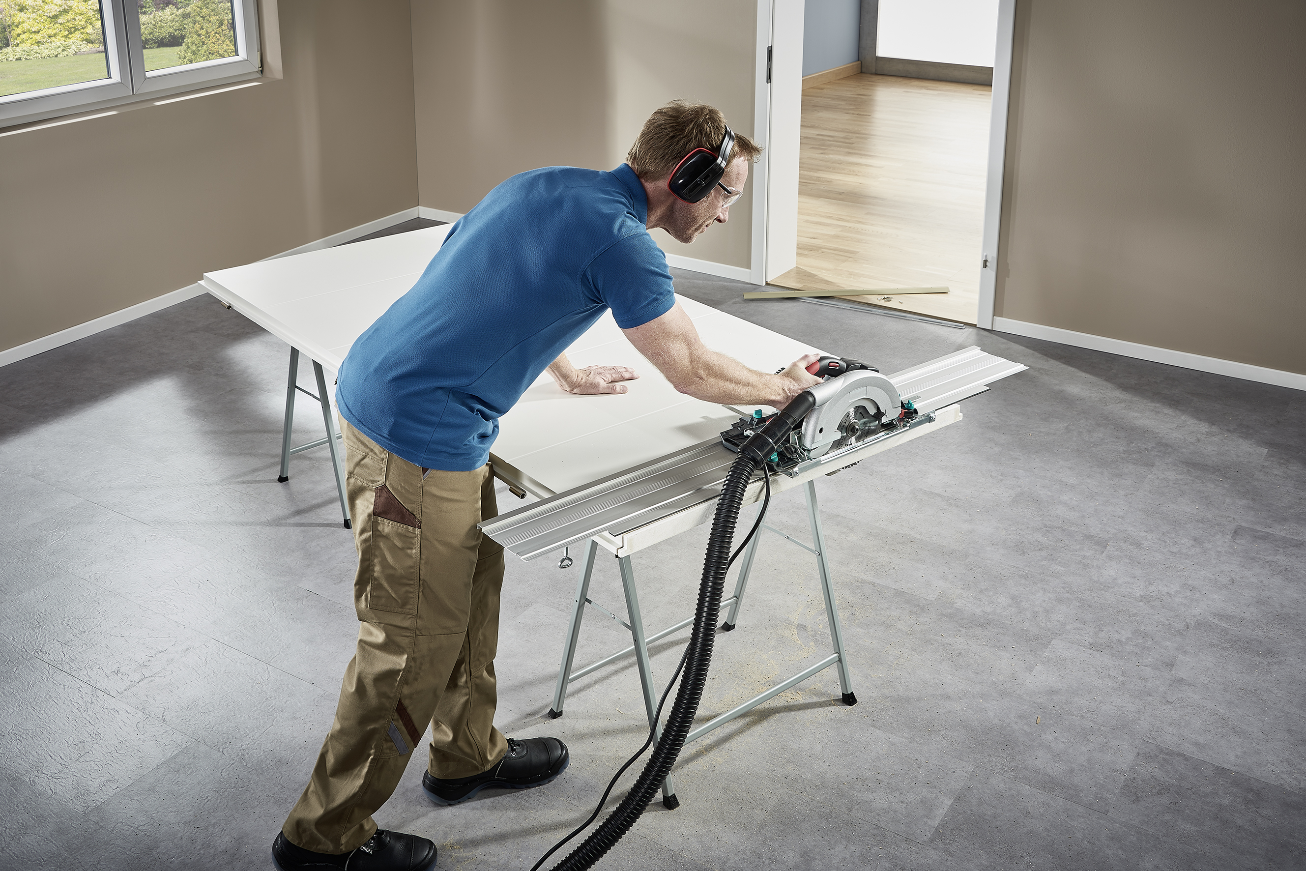 Un homme scie avec une scie circulaire sur une table dans une pièce vide, portant des protections auditives et des vêtements de travail.