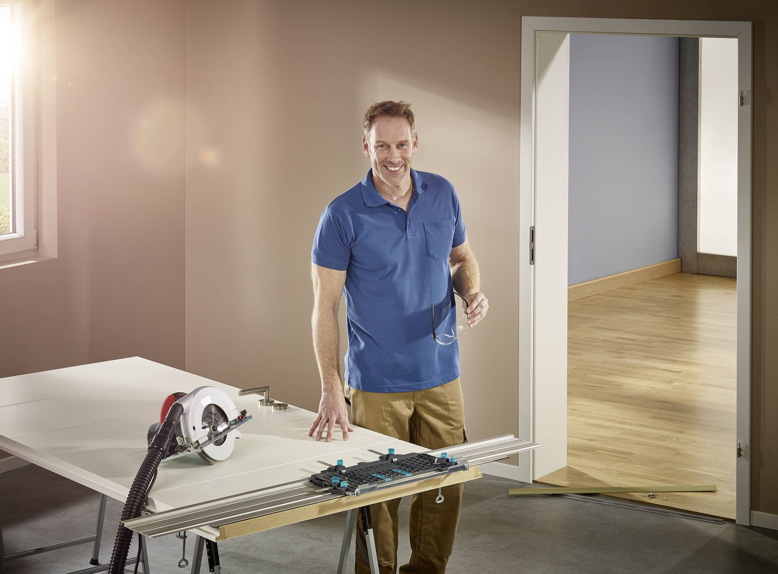 Un homme souriant portant un polo bleu travaille dans une pièce avec une scie circulaire posée sur une table à côté de lui. Il se trouve dans une pièce bien éclairée avec un sol en bois.