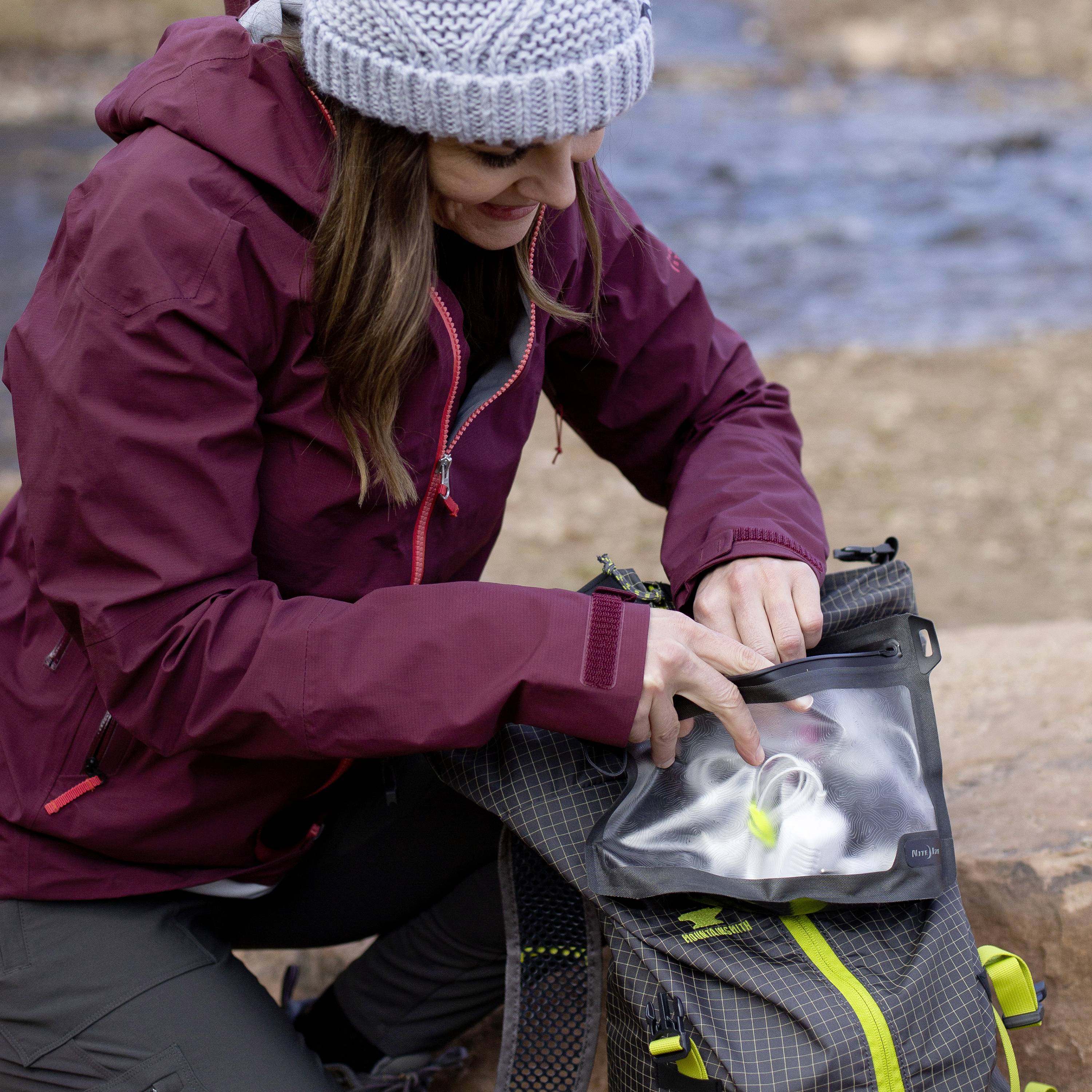 Une personne portant une veste rouge et une casquette grise range des objets dans un sac à dos gris. En arrière-plan : des rochers et un terrain flou.