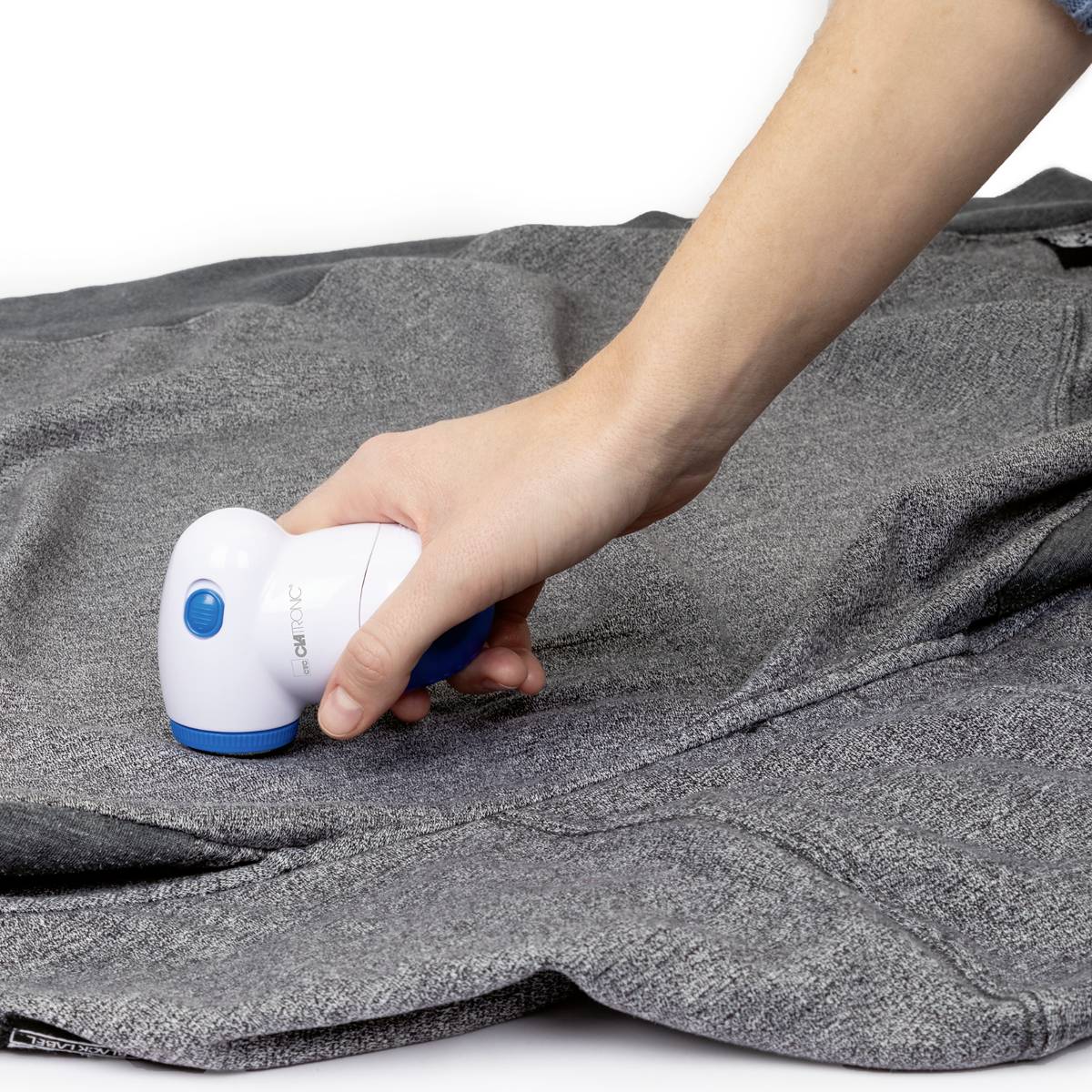 Une main utilise un rasoir anti-peluches électrique sur un tissu gris pour enlever les peluches et les fibres.