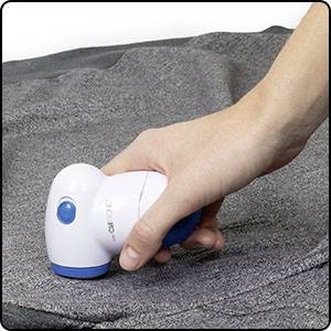 Une main tient un rasoir anti-peluches au-dessus d'un tissu gris pour enlever les peluches. L'appareil est blanc avec un détail bleu.