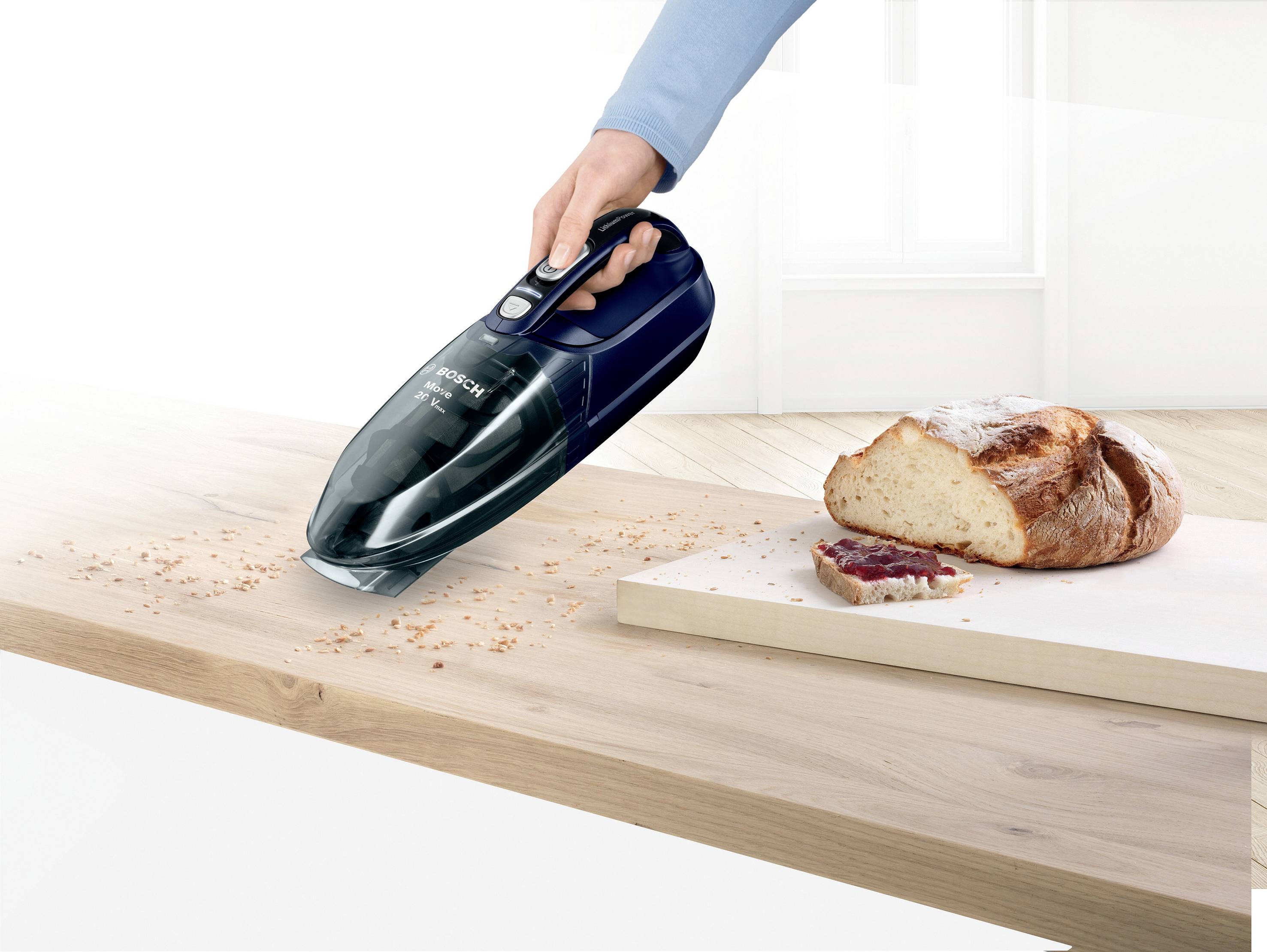 Une main tient un aspirateur à main qui enlève des miettes d'une table. À côté de l'aspirateur se trouve un pain de pain avec une tranche coupée.