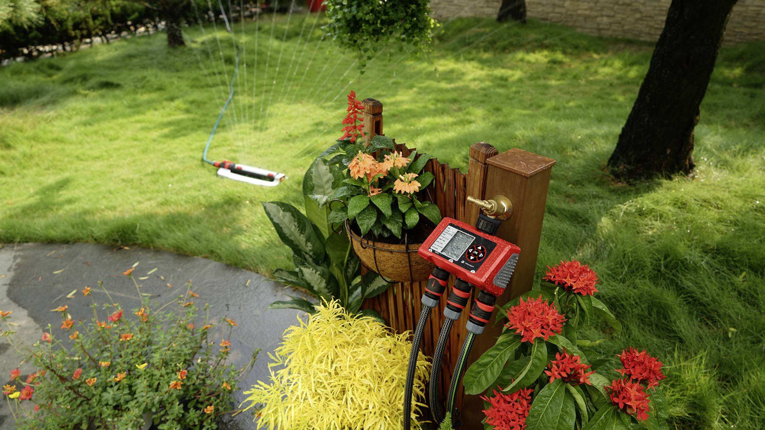Un jardin avec des fleurs colorées, un système d'irrigation équipé de commandes numériques et un arroseur automatique en arrière-plan sur une pelouse.