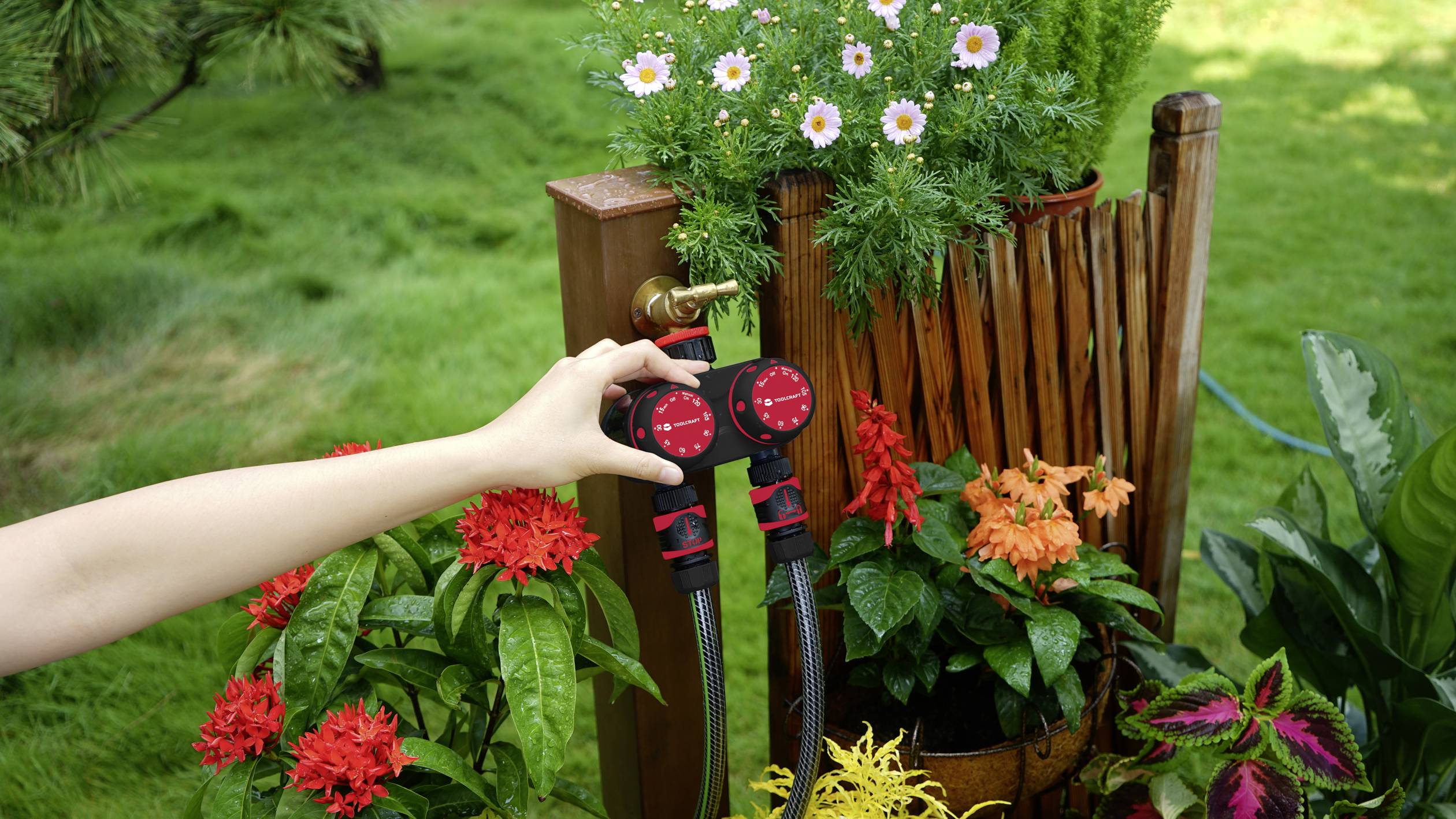 Une personne ajuste deux vannes d'eau rouges sur un robinet de jardin, entourée de plantes en fleurs et d'une pelouse verte en arrière-plan.