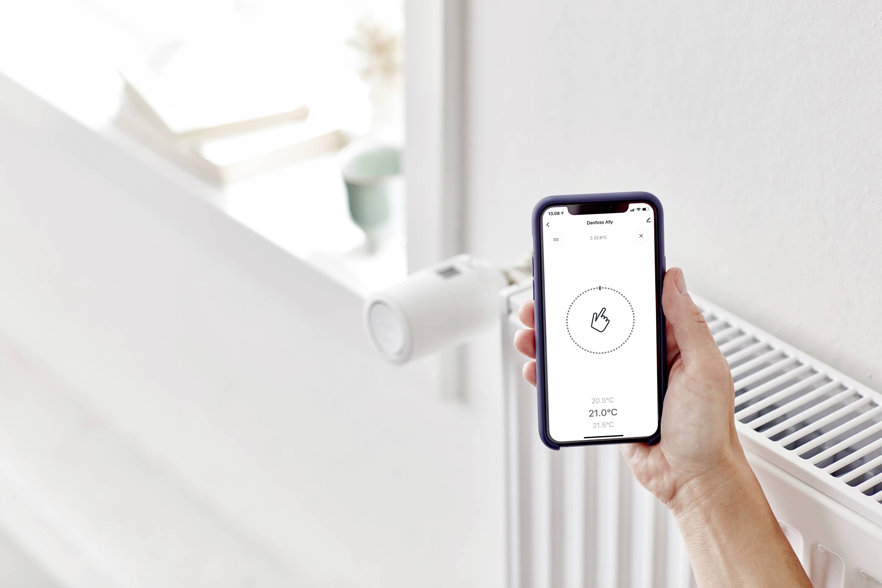 Une main tient un smartphone avec une application de contrôle du chauffage. En arrière-plan, on aperçoit un radiateur équipé d'un thermostat intelligent.