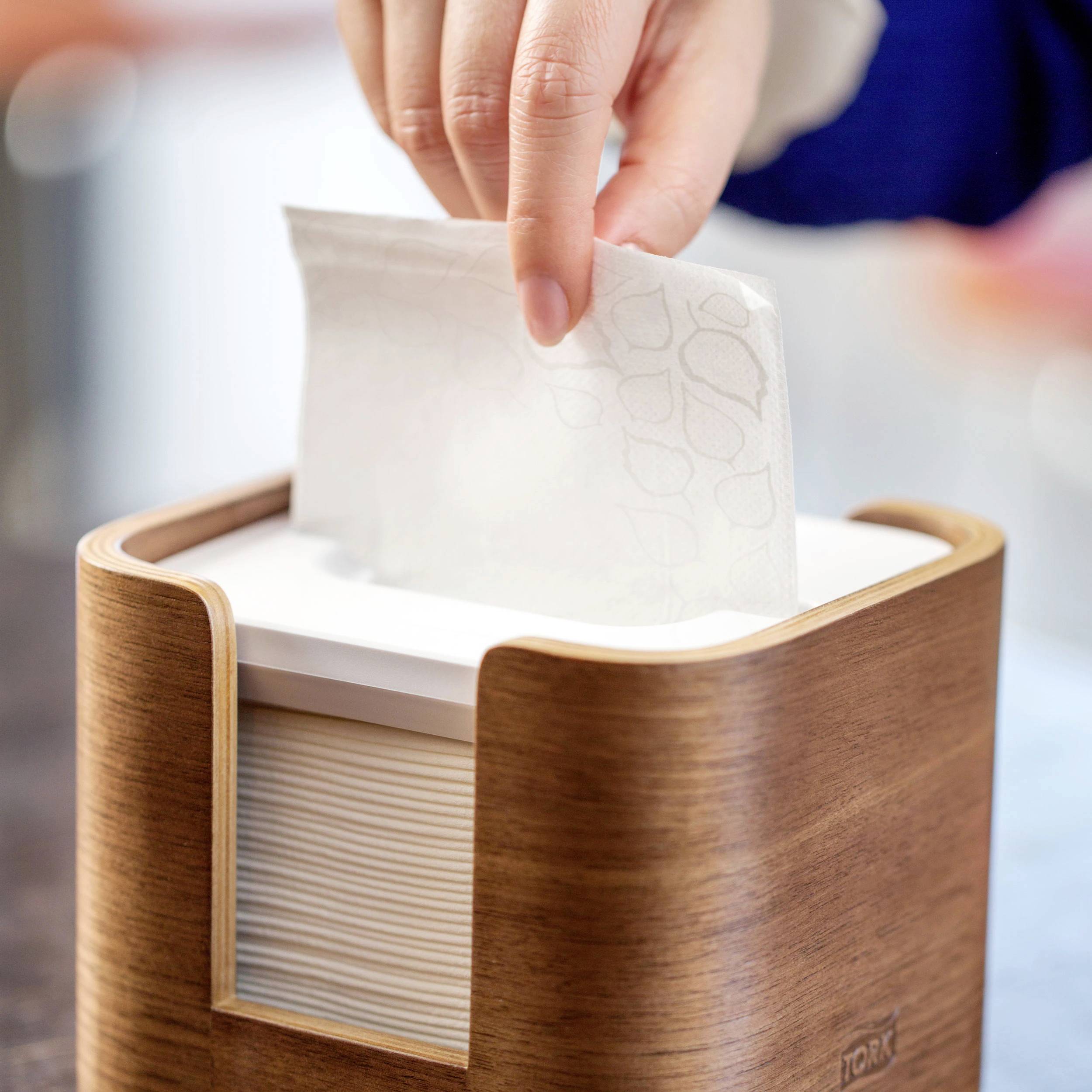 Une main tire un essuie-tout d'un distributeur en bois posé sur une table.