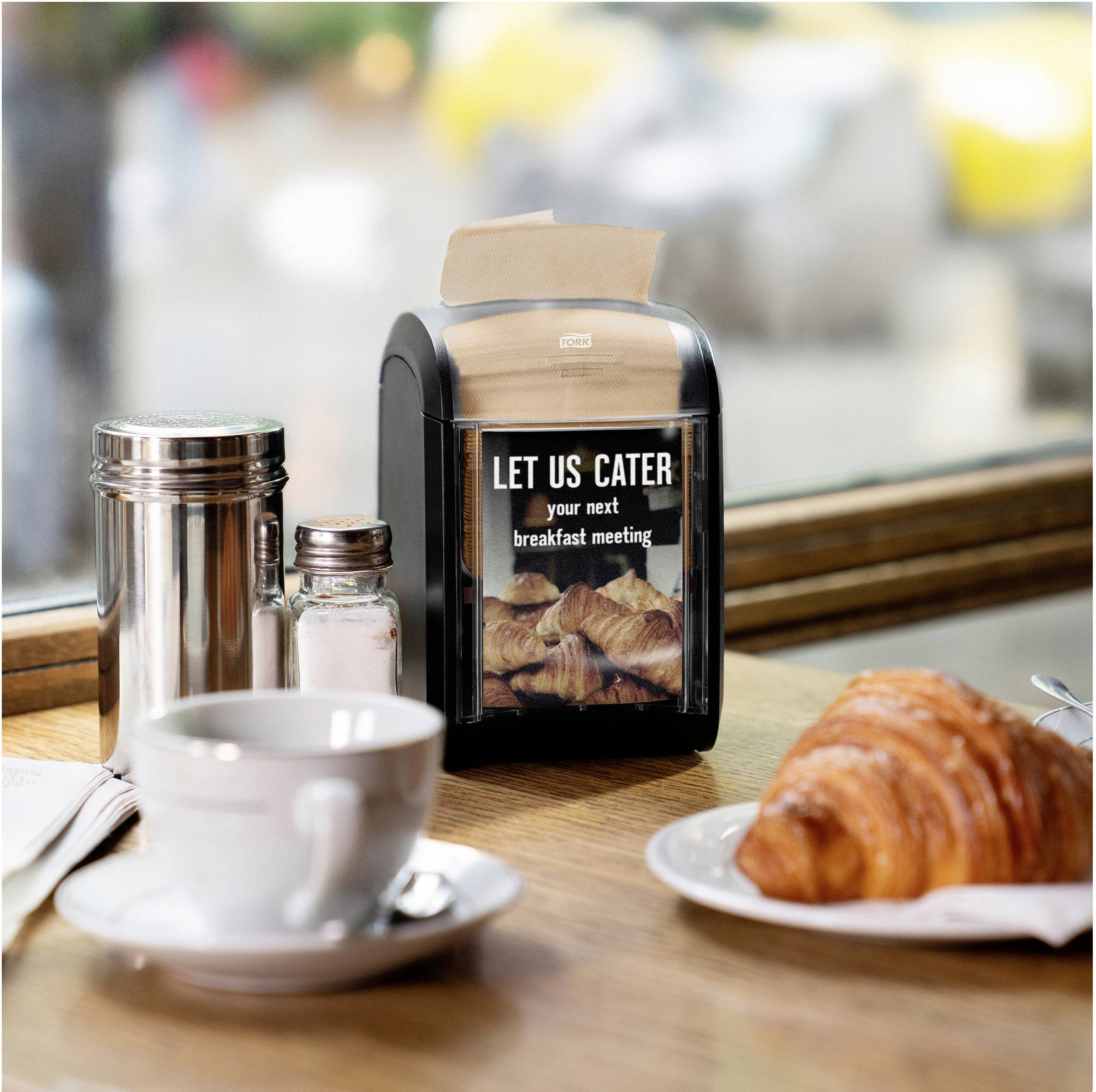 Une table dans un café avec un croissant, une tasse de café, un salière et un sucrier. Au centre, un écran affiche : « LET US CATER your next breakfast meeting ».