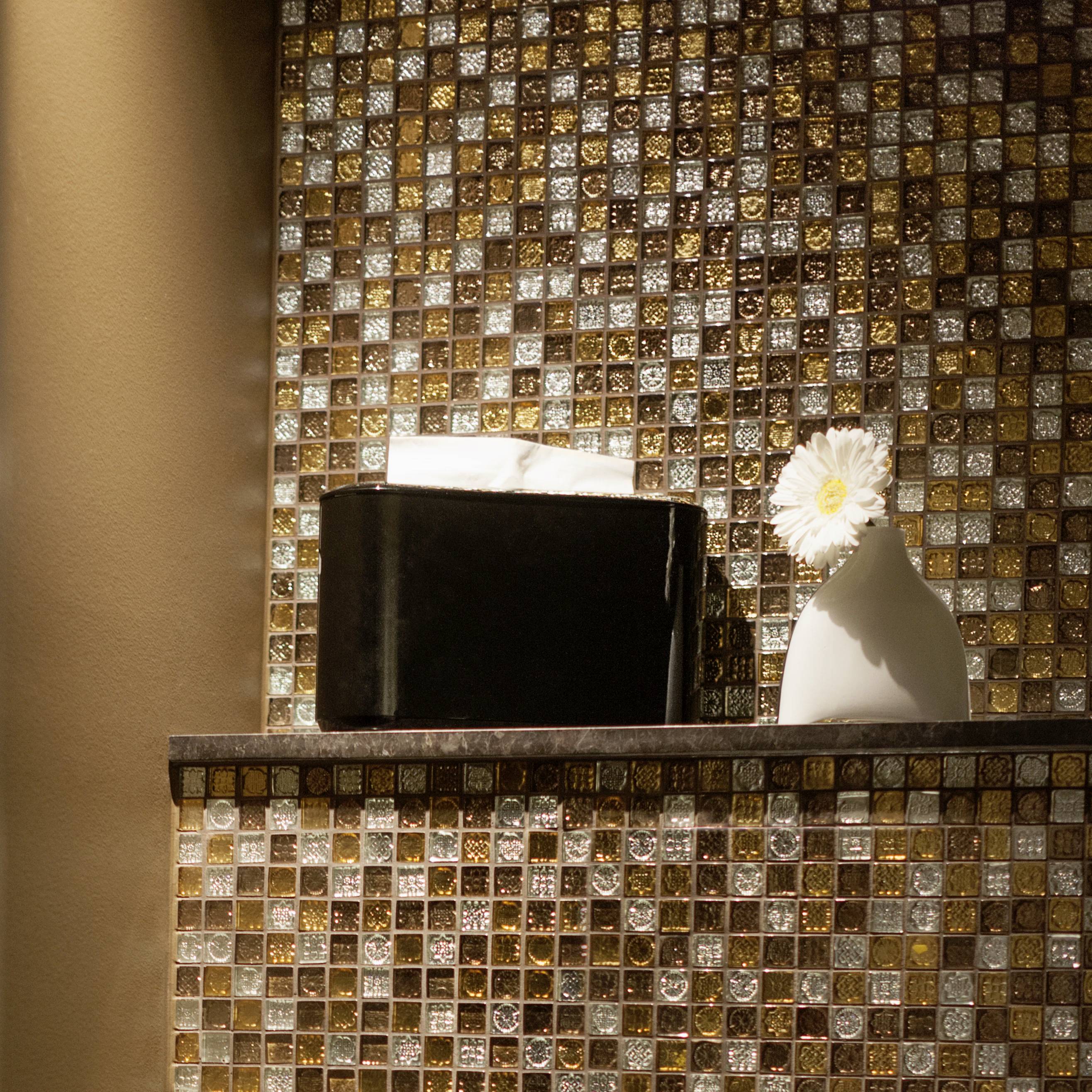 Une étagère avec une boîte noire et un vase blanc contenant une fleur, éclairée devant un mur de mosaïque scintillant.