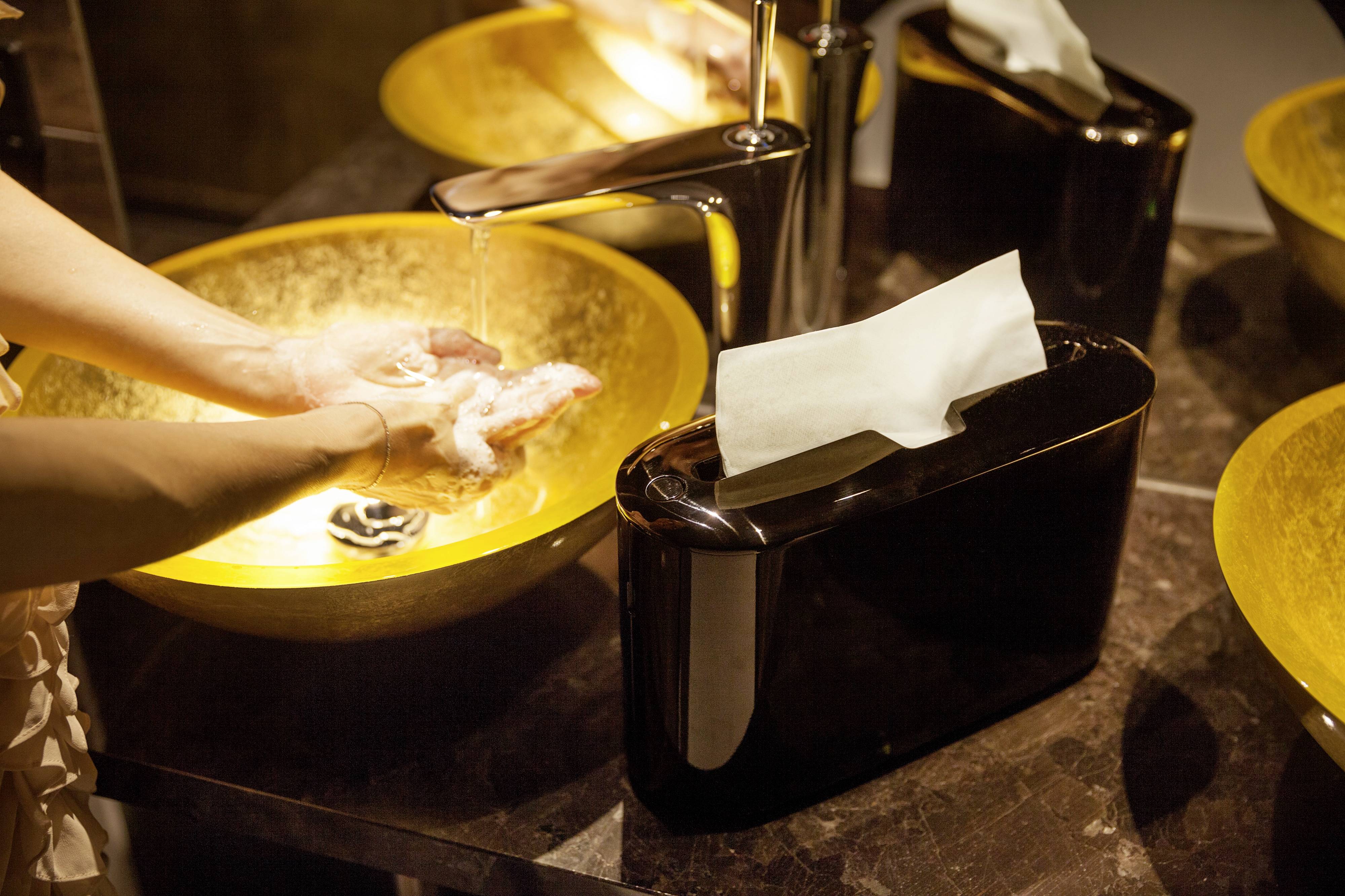 Une personne se lave les mains sous de l'eau courante dans un lavabo doré ; un distributeur noir de serviettes en papier est à côté.