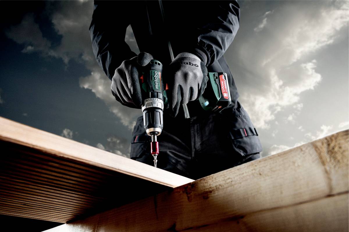 Une personne utilise une visseuse sans fil pour assembler des lattes de bois ; des nuages sombres sont visibles dans le ciel en arrière-plan.