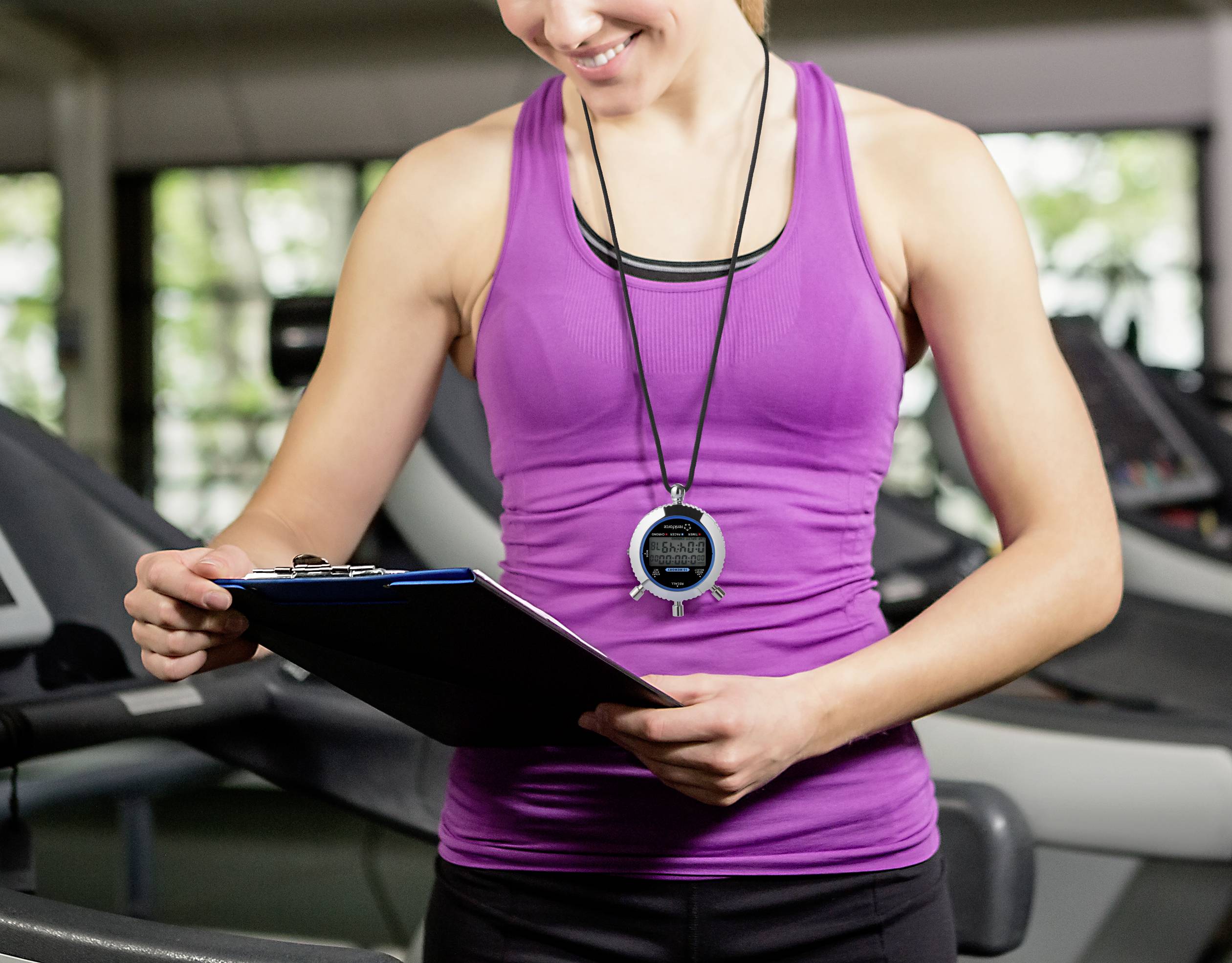 Une personne en tenue de sport, une chronomètre autour du cou, sourit et examine un presse-papiers, probablement dans une salle de fitness.