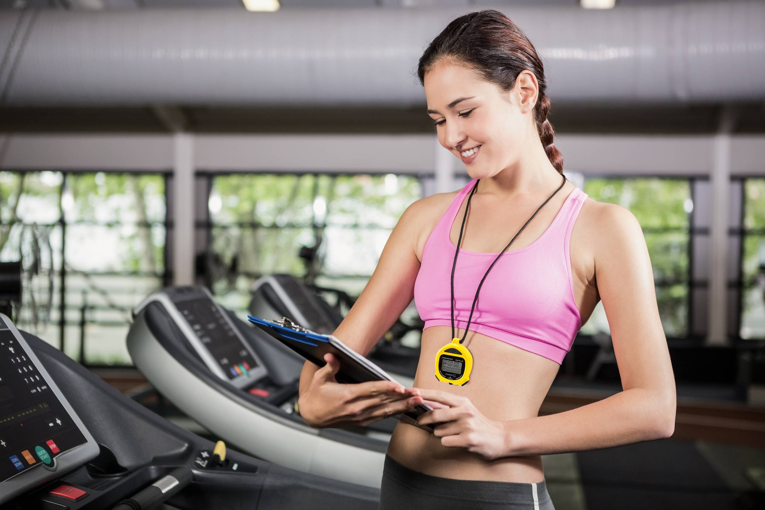 Une femme en tenue de sport se tient dans une salle de fitness, lit quelque chose sur un presse-papiers et porte un chronomètre autour du cou.