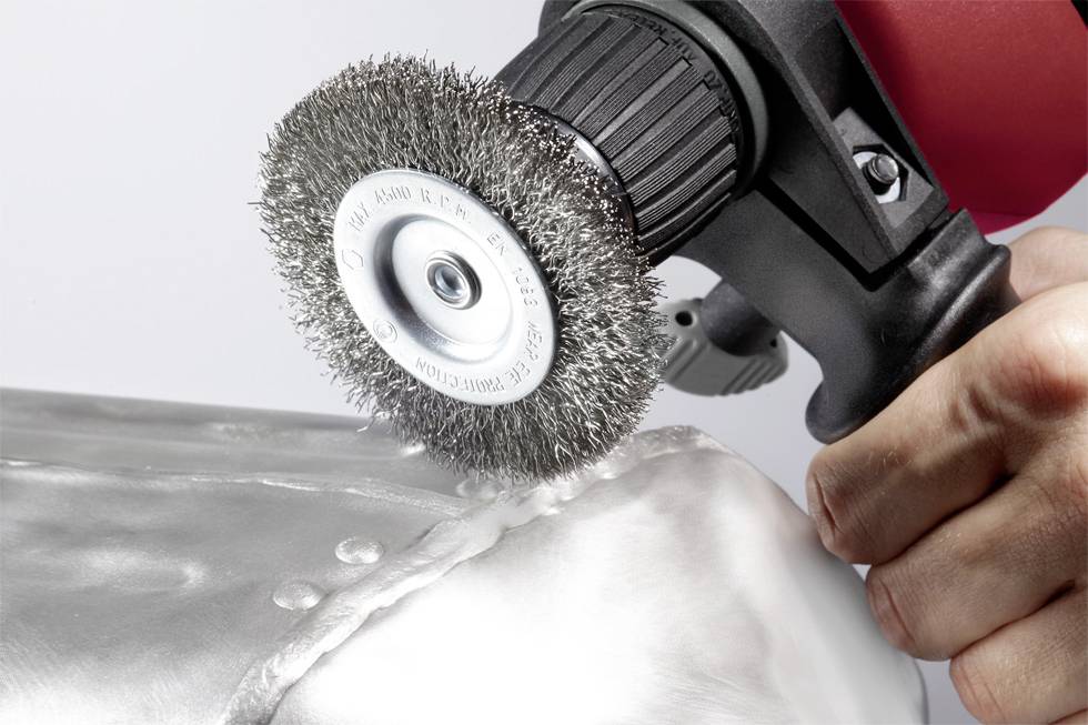 Une main tient un outil électrique équipé d'une brosse métallique, polissant une surface métallique. L'accent est mis sur la manipulation de l'outil.