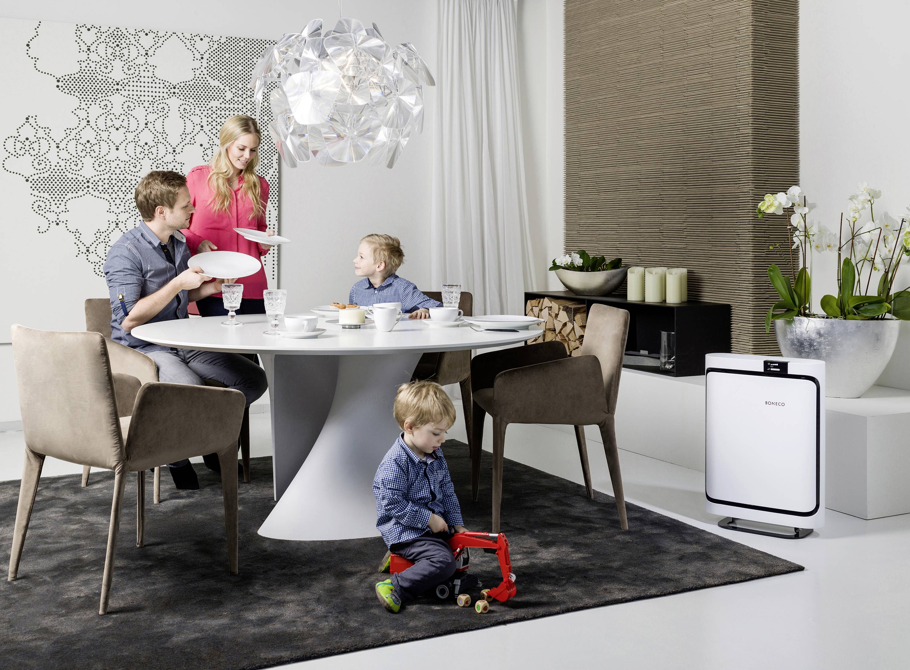 Une famille avec deux enfants prend son petit-déjeuner à une table à manger moderne. Un enfant joue avec un camion-jouet sur le sol.