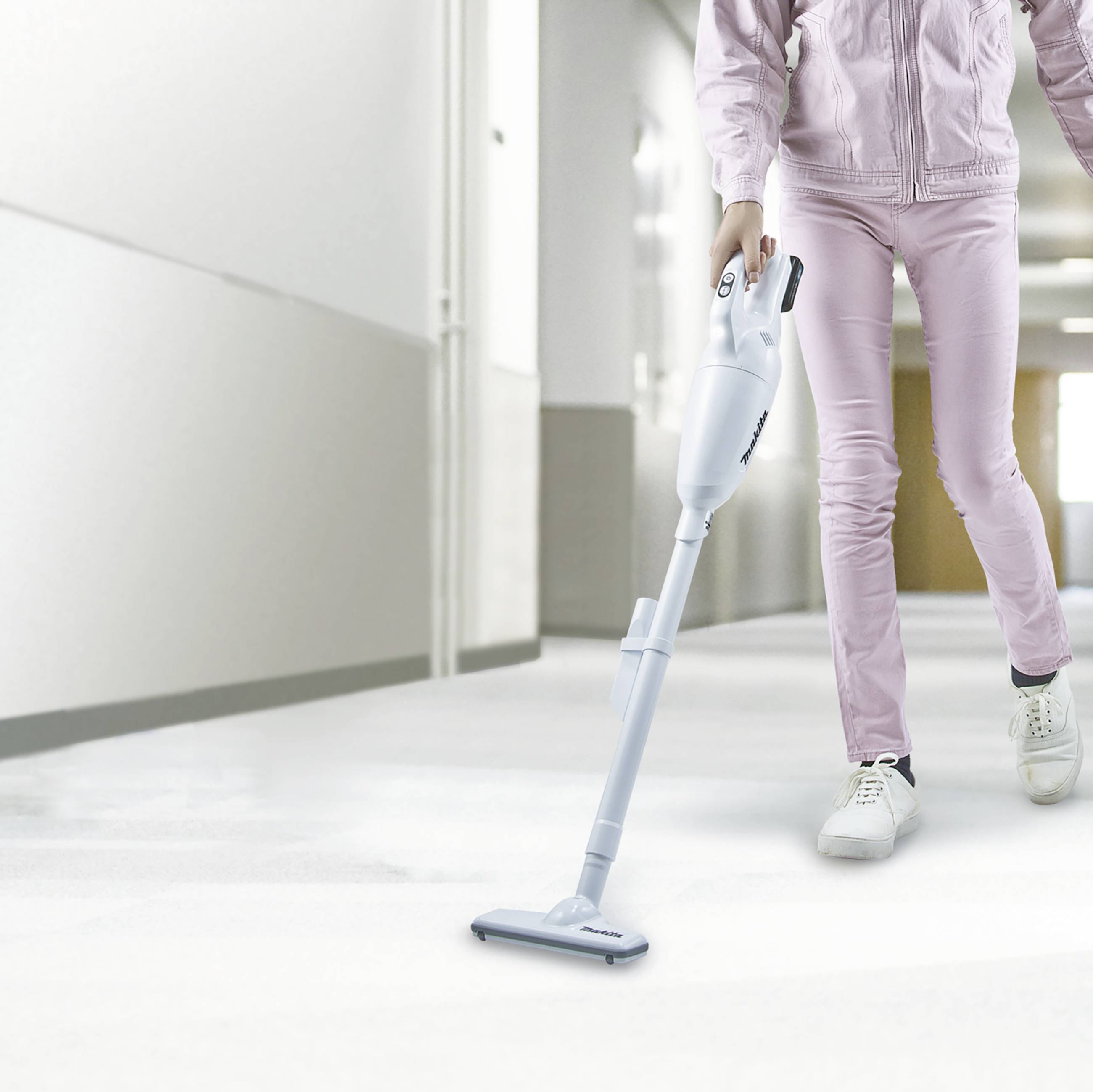Une personne vêtue d'un survêtement rose aspire un couloir lumineux. L'aspirateur est compact et sans fil.