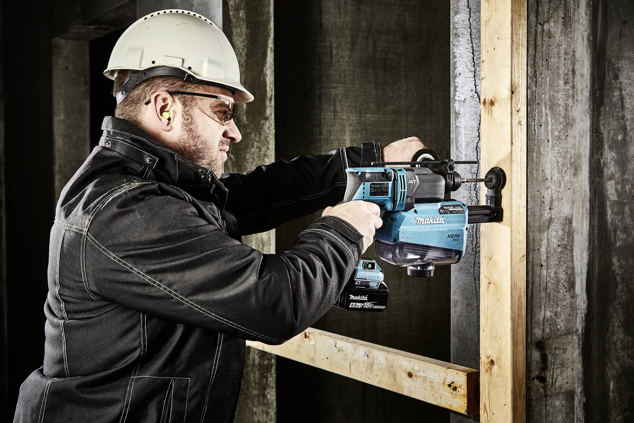 Un ouvrier portant un casque de chantier et des bouchons d'oreilles utilise une perceuse bleue pour percer des trous dans un cadre en bois vertical.