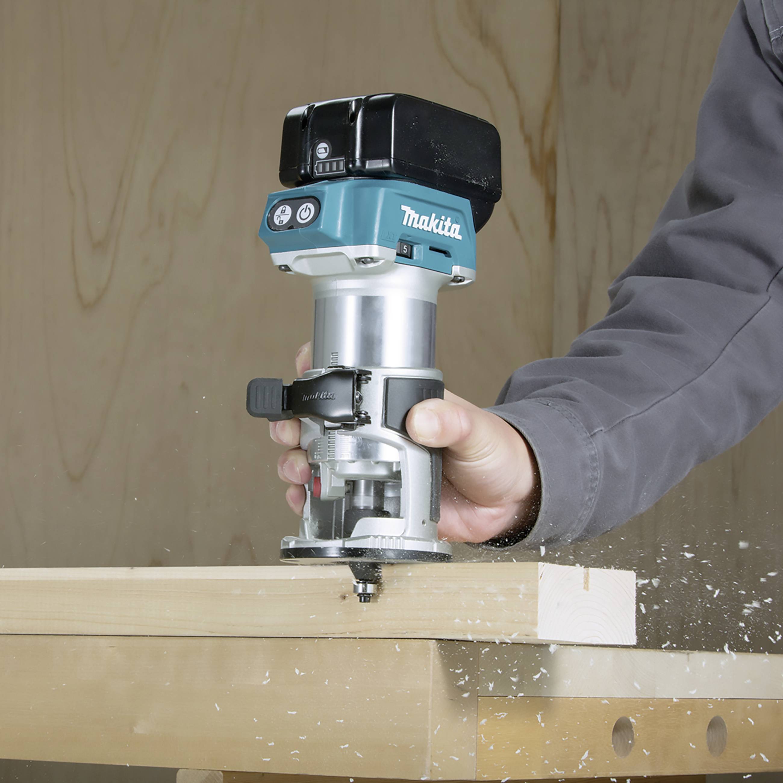 Une personne travaille une planche en bois avec un outil de fraisage bleu Makita, des copeaux volent.